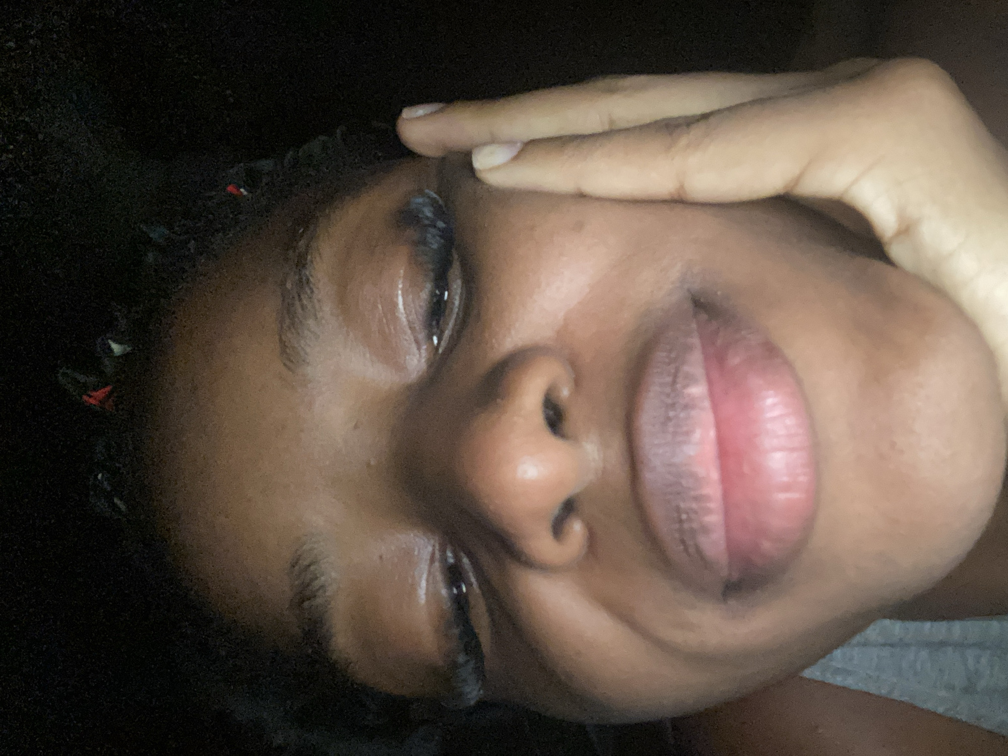 The image is a close-up, low-angle selfie of a young Black person. Their face is partially illuminated, with the lower half of their face, including their mouth and nose, being the most visible. They have full lips with a reddish-pink hue and appear to be wearing false eyelashes. One hand is placed on their cheek, with fingers extended upwards towards their eye. The background is dark and indistinct. There are no discernible landmarks or environmental cues within the image to suggest a specific location in Jos, Nigeria. The image is primarily focused on the individual's facial features and their hand.