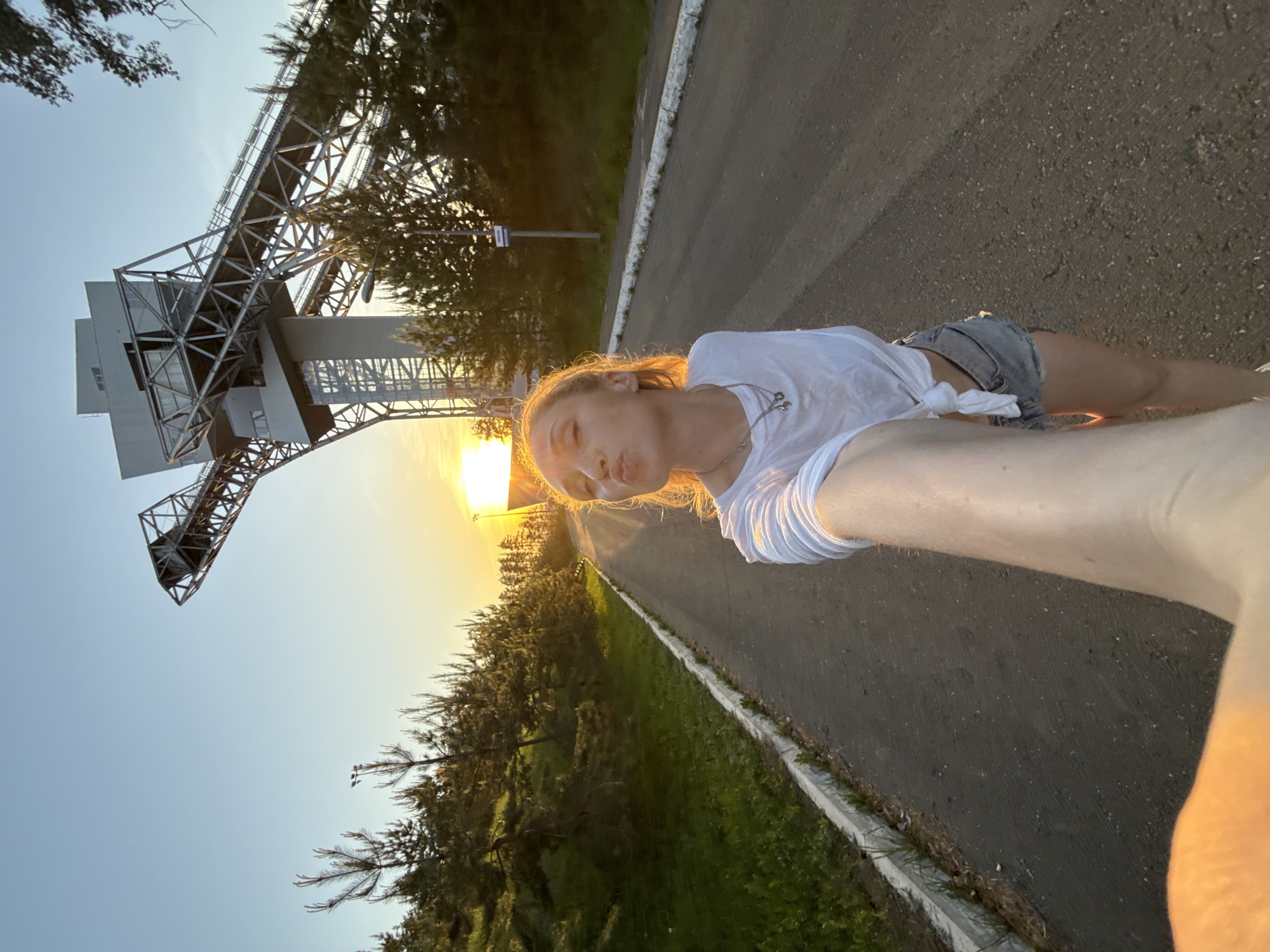 A daytime photograph shows a young woman with blonde hair wearing a white shirt and denim shorts, lying on an asphalt road, with a metal tower structure visible in the background. The tower has a complex framework of steel beams and supports, suggestive of an industrial or transportation structure. Sunlight is filtered through the trees and falls onto the road and the woman, creating a warm glow. Green trees and vegetation are visible along the roadside, suggesting a semi-urban or park setting. The photograph was taken in Прикамский, Russia.