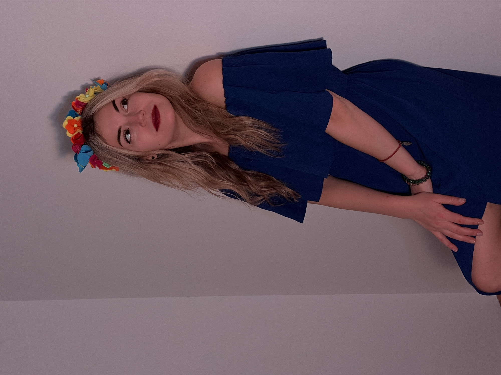A young adult woman is depicted in a portrait, positioned diagonally across the frame against a plain, light-colored background. Her head is tilted upwards, and her body is angled, suggesting she is reclining or leaning against a wall.
She has long, wavy, blonde or light brown hair that flows over her shoulders and chest. Her facial features include full eyebrows, dark eyeliner, and dark red lipstick. Her gaze is directed slightly upwards and to her right, with a thoughtful or serene expression. She wears a vibrant flower crown on her head, made of various fabric flowers in shades of yellow, orange, red, blue, purple, and green.
She is dressed in a dark blue, off-the-shoulder or cold-shoulder dress with wide, flowing sleeves. The dress appears to be made of a light, soft fabric, gathered at the waist, and having a full skirt.
On her right wrist, a thin red string bracelet with a small, light-colored metallic charm (possibly a feather or leaf) is visible. On her left wrist, she wears a bracelet made of dark green, rounded beads. Her left hand is gently placed on her lap or upper thigh, resting on the fabric of her dress.
The background is a simple, unadorned light gray or off-white wall. A subtle horizontal line, possibly a corner or a baseboard, is visible across the lower portion of the background, creating a slight variation in shade. The lighting is soft and even, suggesting an indoor setting. There is no visible text in the image.