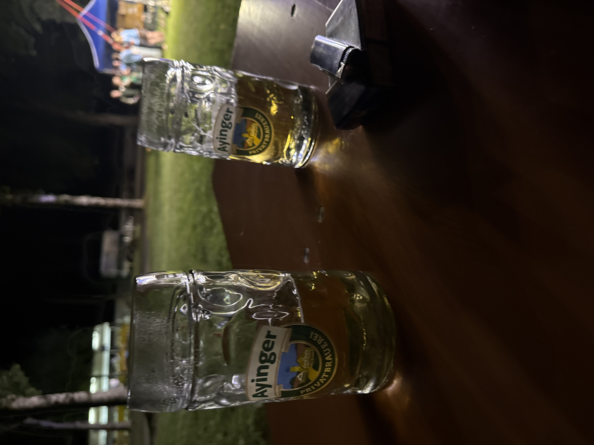 Two beer steins with the Ayinger brewery logo are on a wooden table. The stein on the left is full of beer, while the one on the right is about half full. To the right of the steins, a silver lighter and a small stack of papers are visible on the table. In the background, blurred figures are seen near a blue and red tent, and trees are visible against a dark sky. The scene appears to be an outdoor event, likely at dusk or at night, in the municipality of Höhenkirchen-Siegertsbrunn, Germany.