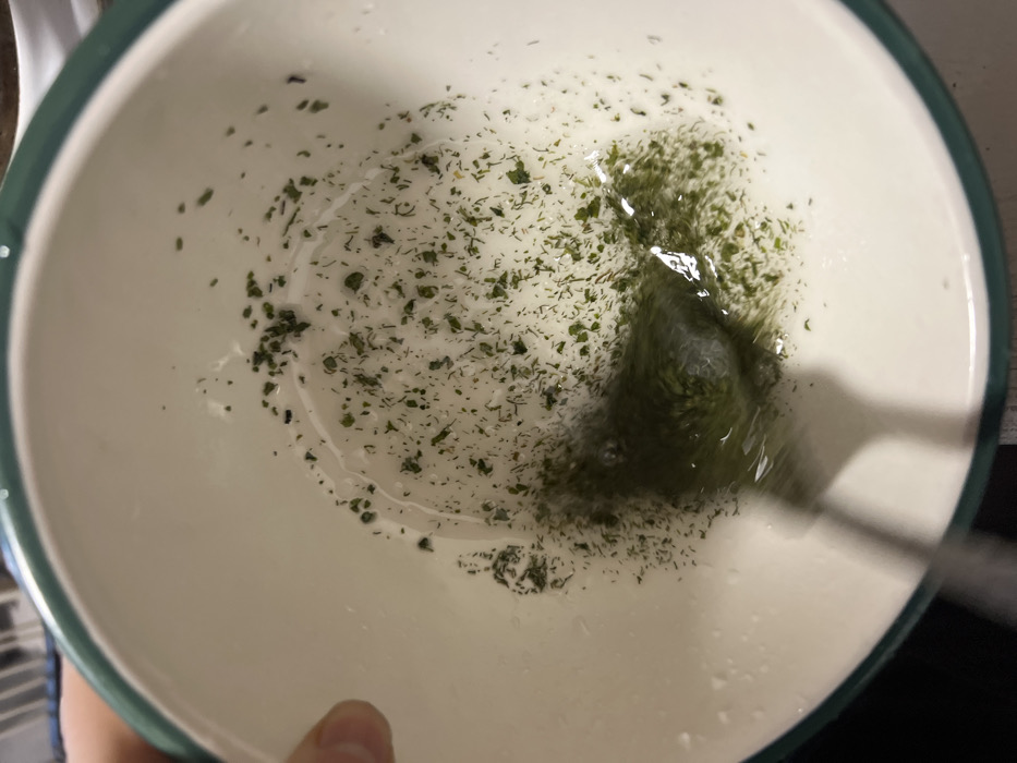 An overhead photograph depicts a white ceramic bowl featuring a dark green rim. The bowl contains a shallow, clear liquid in which numerous small, dark green fragments, consistent with dried herbs, are dispersed. A metallic utensil, blurred by motion, is actively stirring the liquid and herbs in the lower-center of the bowl, creating a localized swirl and denser concentration of the green particles. A partial human hand, with a visible thumb and part of an index finger, holds the upper-left edge of the bowl. The background outside the bowl is out of focus and indistinct. There are no visual elements within the image to identify the specific location as Saint Petersburg, Russia; the scene appears to be an indoor setting, likely a kitchen.