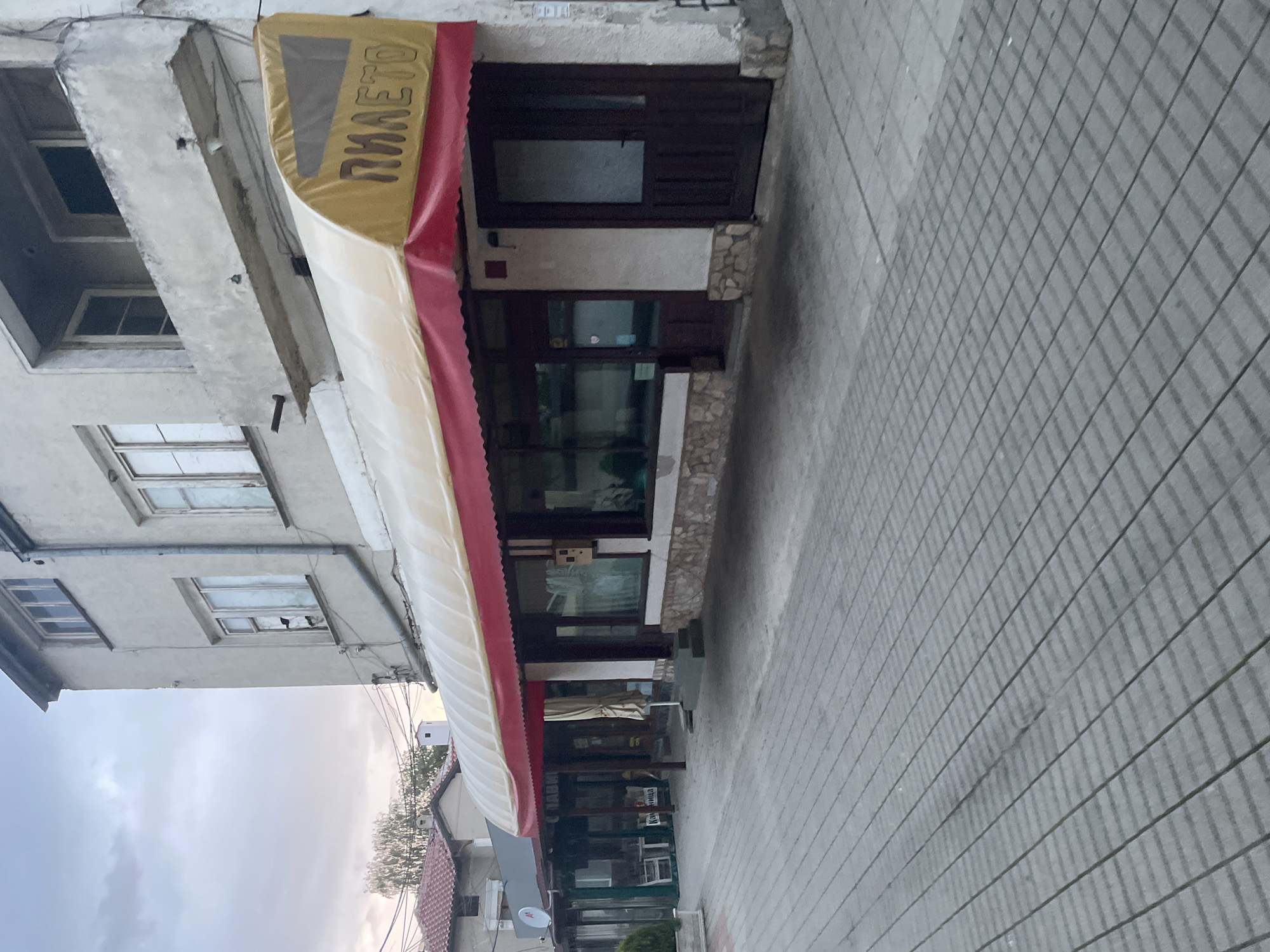 This is an exterior photograph of a street scene in Ihtiman, Bulgaria, showing the ground floor of an older building with a shopfront and an awning, alongside a tiled sidewalk.
The main subject is a shop with a prominent awning. The building itself appears to be made of concrete, possibly aged, with several windows visible on the upper floors, some featuring white frames. The shop entrance on the ground floor has a dark wooden door and large glass windows. The facade around the shopfront is decorated with irregular stone cladding.
An awning extends over the shopfront. Its top surface is golden yellow, with a red trim along its edge, and its underside is white. On the yellow part of the awning, the Cyrillic word "ПИЛЕTO" is visible in embossed brown letters, which translates to "PILETO" (meaning "Chicken" in Bulgarian). This suggests the establishment might be a chicken shop, a grill, or a fast-food restaurant specializing in poultry.
To the right of the building, a wide sidewalk is visible, paved with grey square tiles arranged in a grid pattern. The sky in the background appears overcast or cloudy, and hints of distant buildings and trees can be seen.
The lighting indicates it is daytime, possibly an overcast day or late afternoon, as shadows are subtle. No people or specific activities are occurring in the visible portion of the street. The scene is static, capturing a quiet moment in a Bulgarian town.