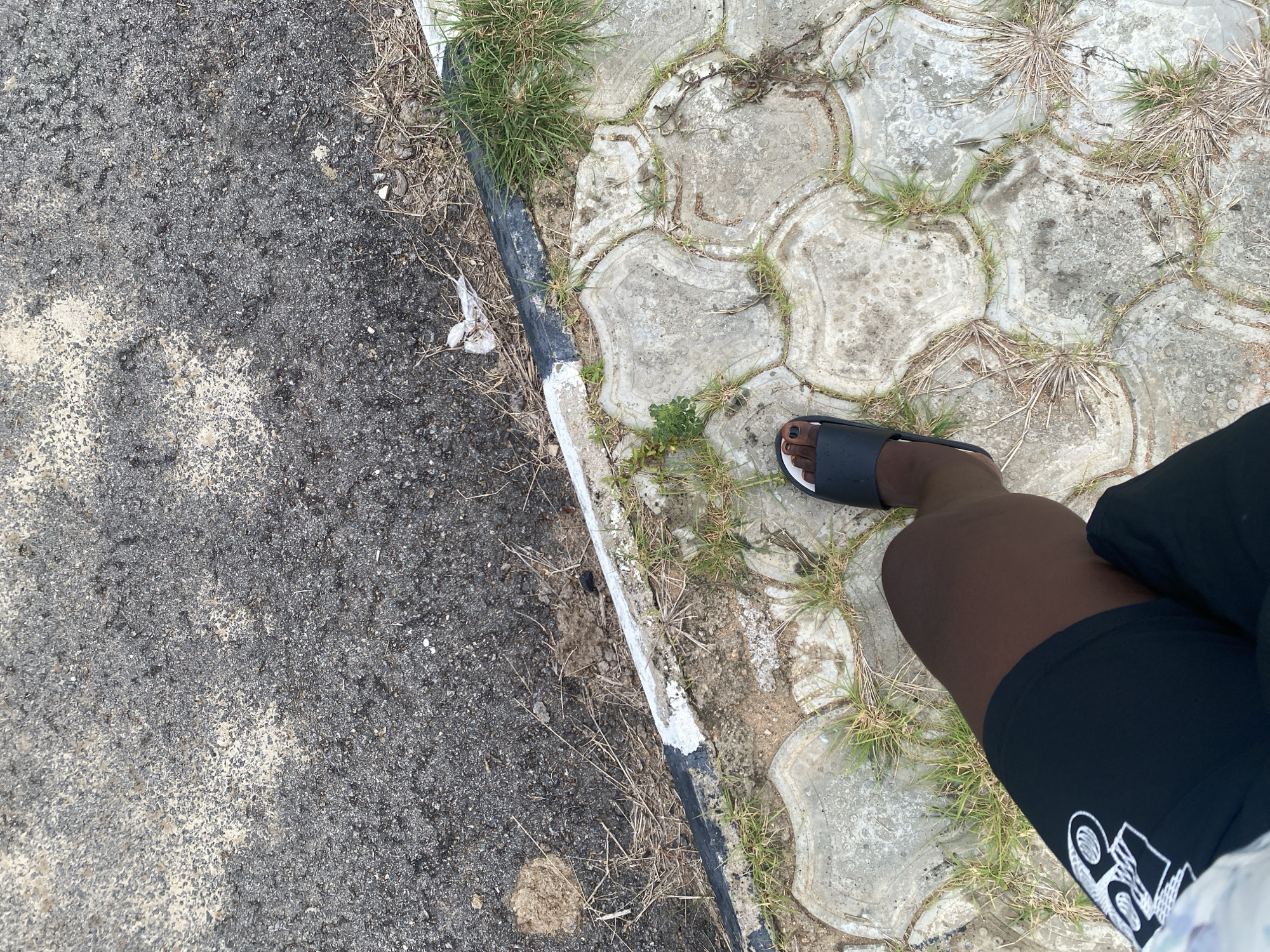 The image shows a person's legs and feet standing on a tiled pathway next to an asphalt road. The pathway is made of interlocking concrete tiles with grass growing between them. The asphalt road is textured and appears worn. A curb with a black and white line separates the road from the pathway. The scene is outdoors, and the lighting suggests it is daytime. There is some dry grass and debris visible on the edge of the road. The person is wearing black shorts with a white graphic design and black slide sandals. The photograph is taken from a high-angle perspective, looking down at the ground and the person's legs. The location is specified as Dumnga, Nigeria.