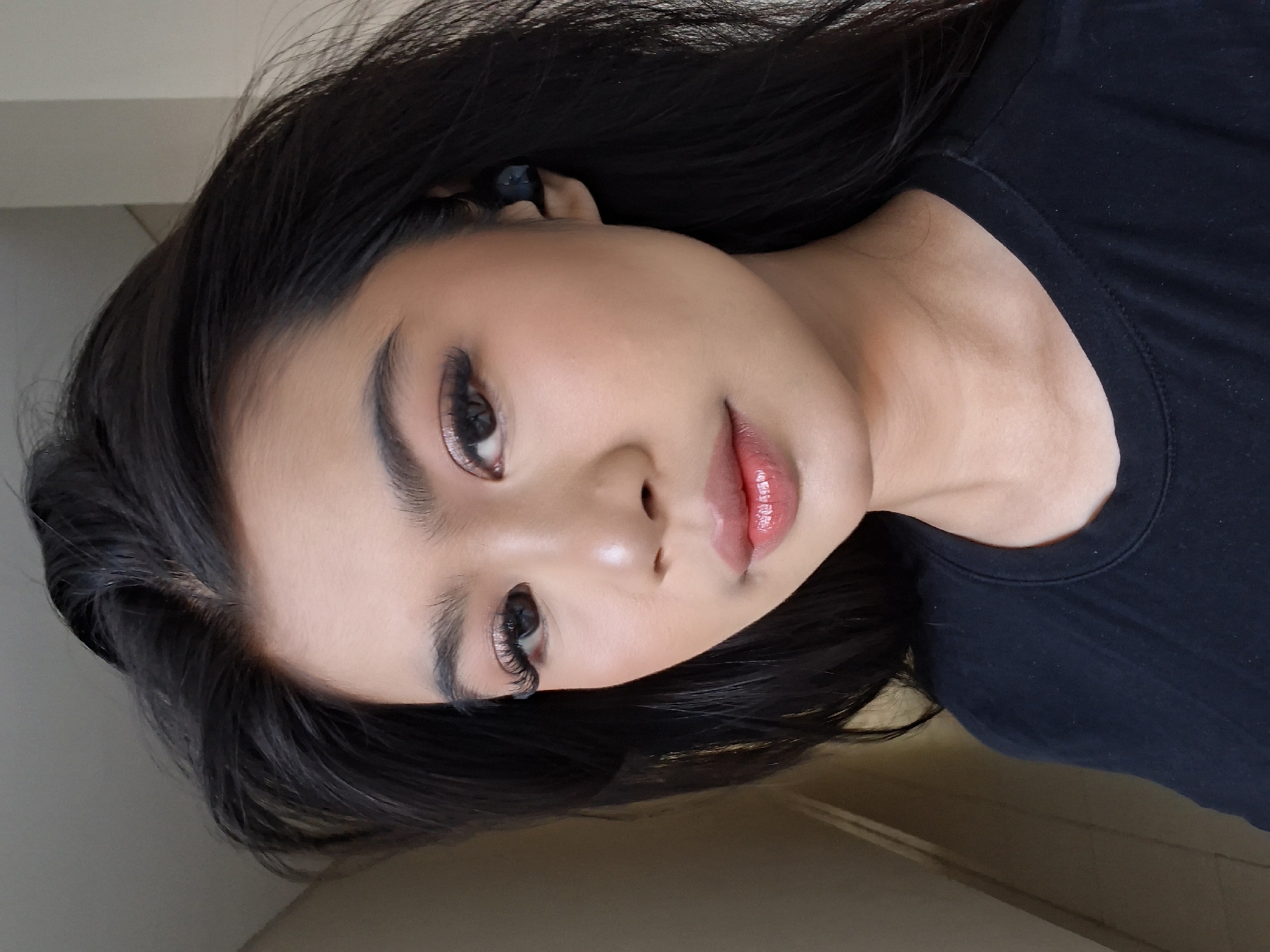 A medium shot shows a young woman from the chest up, looking into the camera. She has dark, wavy hair that frames her face and shoulders. She is wearing a black t-shirt and has a black wireless earbud in her right ear. Her makeup includes foundation, blush, eyeshadow, mascara, and lipstick. The background is a plain, light-colored wall with a hint of a doorway on the left. The lighting appears to be natural, coming from the right side of the frame. The woman's expression is neutral, and her gaze is direct.