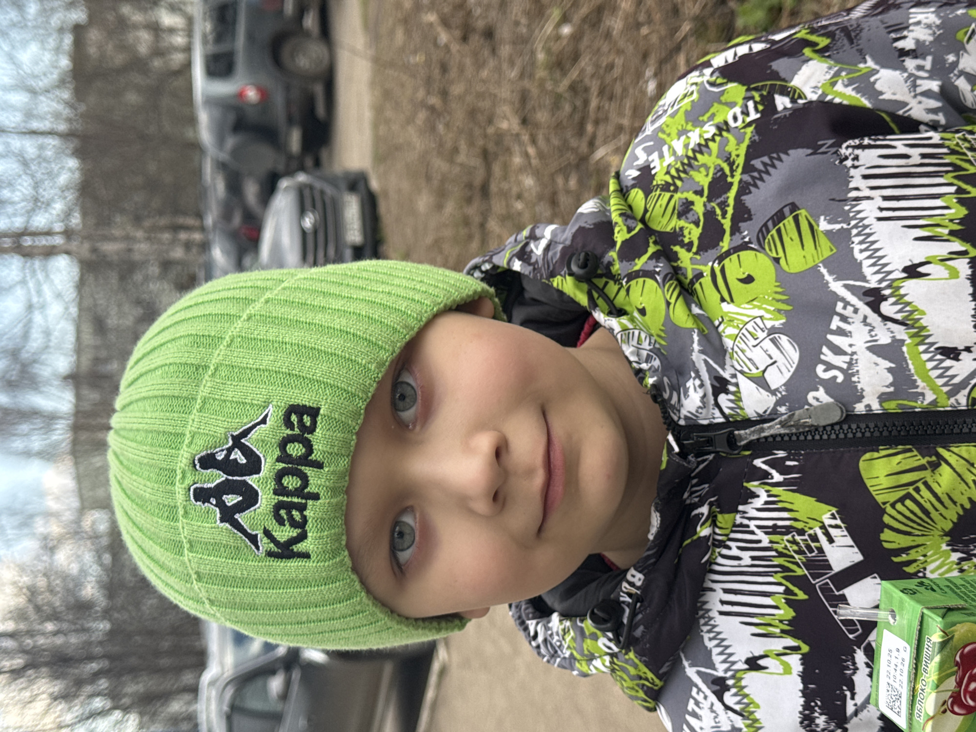 The image displays a close-up outdoor portrait of a young child, likely a boy, rotated 90 degrees counter-clockwise. The child looks directly at the viewer with wide, light-colored eyes and a soft, gentle smile.
He is dressed in a bright lime green ribbed beanie hat, prominently featuring a black Kappa logo and "Kappa" text. He also wears a multi-colored winter jacket with a distinct pattern of black, grey, and neon green. The jacket is adorned with skate-themed graphics and text, including "SKATES" and "SKATEBOARD".
The background is softly blurred, showing an outdoor setting with parked cars and what appear to be bare trees. This suggests a cool climate or the transitional seasons of early spring or late autumn. The lighting indicates it is daytime, possibly under an overcast sky.
A small green juice box with a straw is visible in the lower right corner, partially tucked into the child's jacket. The label on the juice box clearly shows Cyrillic text "ЯБЛОКО-ВИШНЯ" (Apple-Cherry), along with numerical codes such as "22.10.25", "10:44.1.9", and "22.10.26", likely indicating dates or product information. The scene suggests a typical urban or suburban environment in Chaikovsky, Russia. The child's expression conveys a sense of calm and contentment.