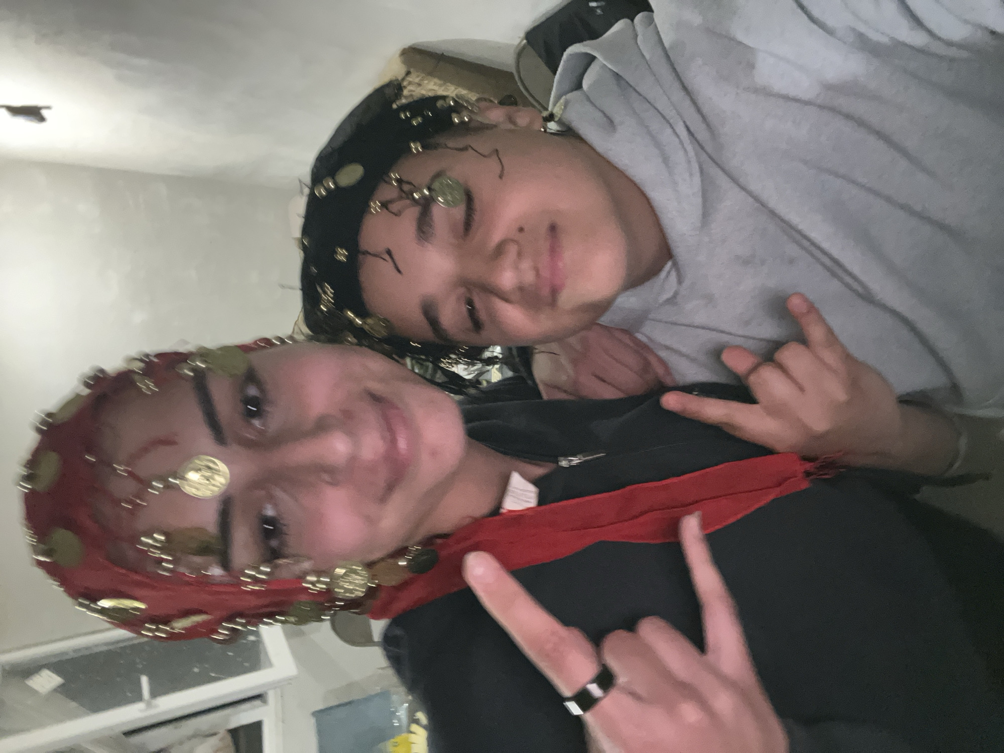 Two individuals pose for a selfie indoors. The foreground person wears a red headscarf adorned with gold coin-like embellishments, a black top with a red scarf around the neck, and a black ring on one finger. They are making a rock-on hand gesture. Behind them, the other individual wears a black headscarf with gold coin-like embellishments and a gray hooded sweatshirt, also making a rock-on hand gesture. The indoor environment includes a window and a white ceiling. There is no indication of Lozarevo, Bulgaria, in the image.