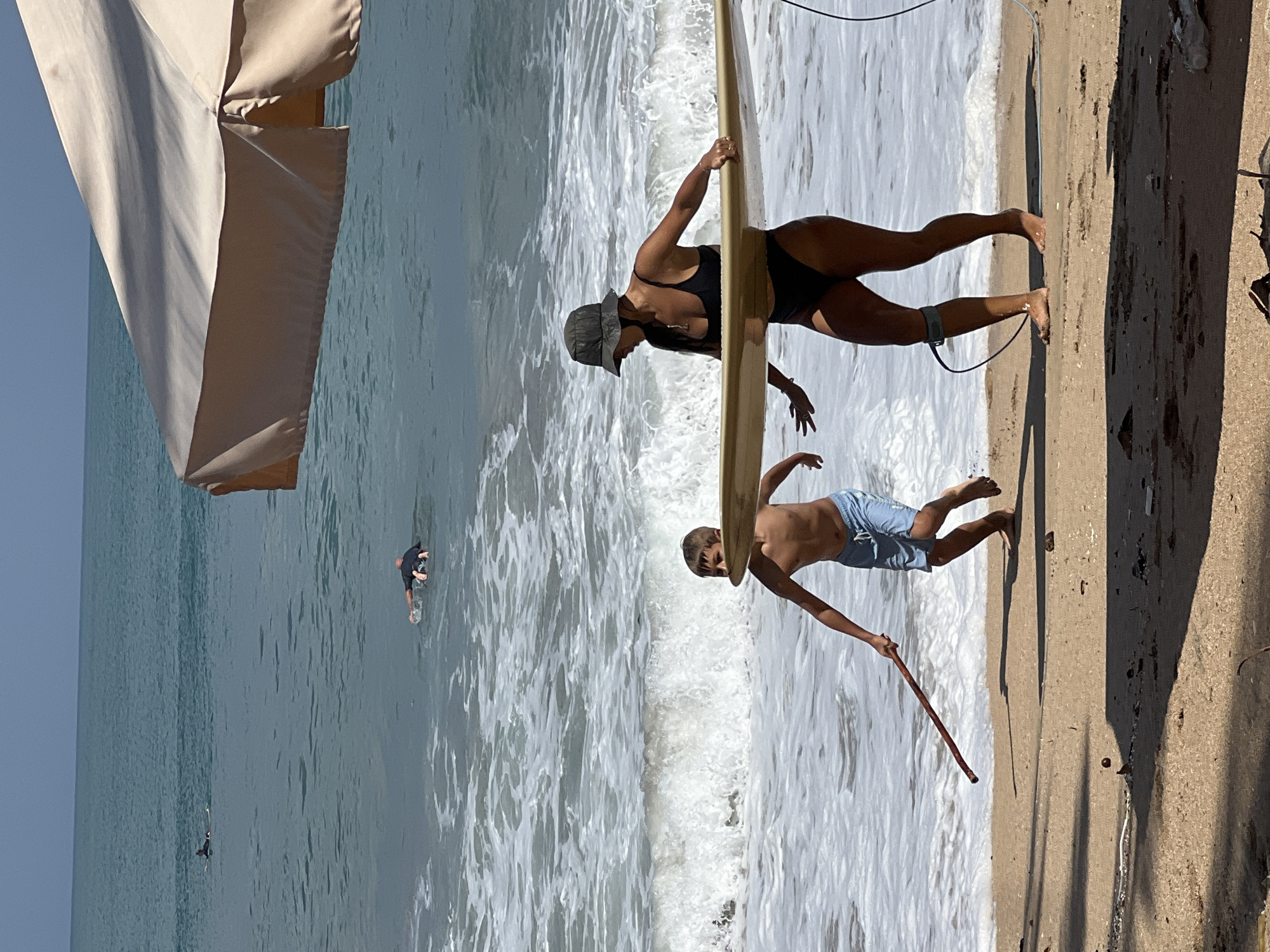 In Kuta, Indonesia, on a sandy beach adjacent to the ocean, a woman wearing a black bikini and a bucket hat is holding a surfboard. She is partially in the water, with waves breaking around her. A young boy, wearing blue swim trunks, is running towards the right of the frame, holding a stick. In the background, several surfers are in the ocean, some on their boards and others paddling. The sky is clear and blue, and a large, tan beach umbrella is visible in the upper right corner of the image.