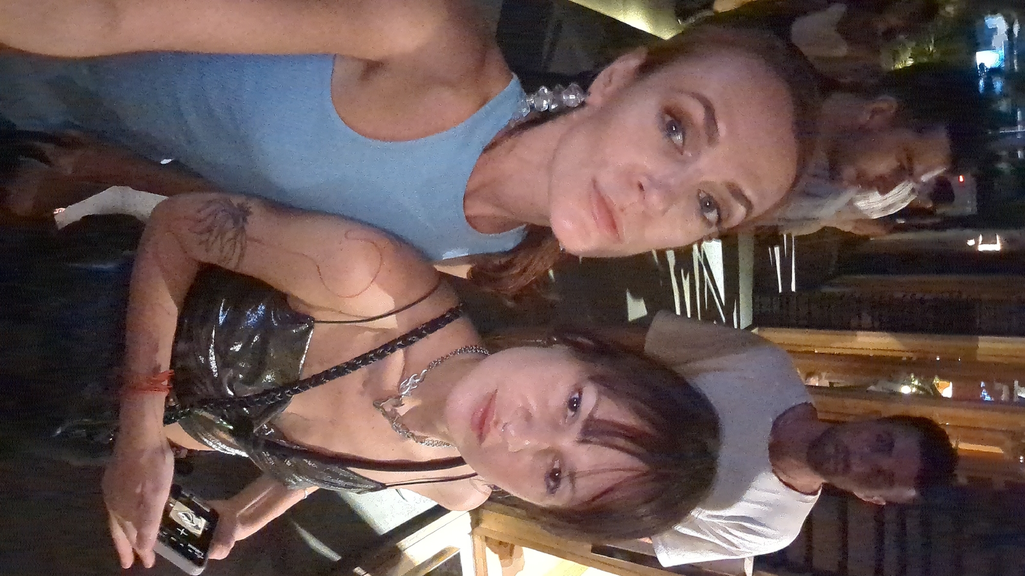 The image, taken at night in North Kuta, Indonesia, features two women posing for a photo, likely a selfie. In the foreground, one woman with light brown hair and blue eyes wears a light blue sleeveless top and large, clear beaded or crystalline earrings. Her expression is neutral. Beside her, another woman with dark hair and bangs wears a shiny black sleeveless top and a silver chain necklace with a distinctive pendant. This woman has a tree-like tattoo on her upper left arm and a reddish bracelet on her left wrist. She is holding a smartphone in her left hand, with partial text "Tra..." visible on its screen. Her expression is also neutral, with a slight smile. Both women are looking directly towards the camera. In the blurred background, several other people, primarily men, are visible, including one in a white t-shirt, suggesting a social setting like a restaurant or bar. The dim lighting indicates it is nighttime.