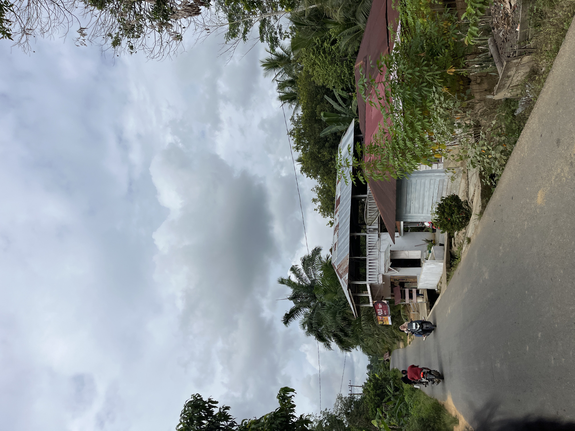 The image depicts a street scene in the city of Sukamulia, Indonesia, under a cloudy sky. A paved road runs through the center of the frame, with lush green vegetation and trees on both sides. Several houses are visible along the road. One house, in the mid-ground, features a white exterior with a balcony and a corrugated metal roof. Adjacent to it is a smaller structure with a red metal roof and light blue wooden siding. In the foreground, two motorcycles are traveling on the road. The first motorcycle, closer to the viewer, is ridden by a person wearing a red shirt. The second motorcycle, further ahead, is ridden by a person wearing a hijab and blue clothing. A few scattered pieces of debris are visible on the side of the road. The overall atmosphere is calm and rural.