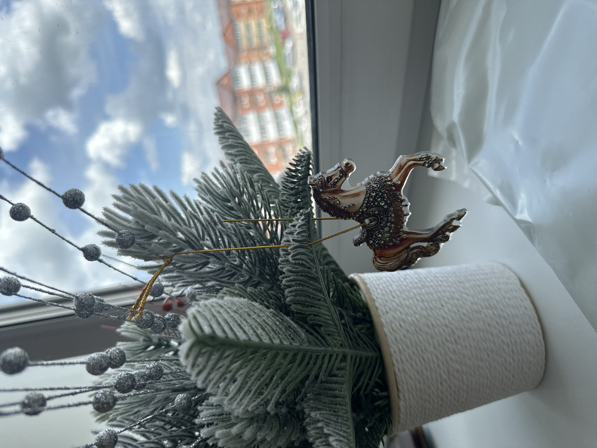 A decorative artificial Christmas tree with a carousel horse ornament is positioned on a windowsill. The tree is white and appears to be frosted. The ornament, a carousel horse, is transparent with a metallic sheen and intricate details, hanging from a gold string. In the background, a window shows a cloudy sky and apartment buildings in Ussurijsk, Russia. The foreground includes a white textured pot holding the tree and a white, wrinkled cloth.