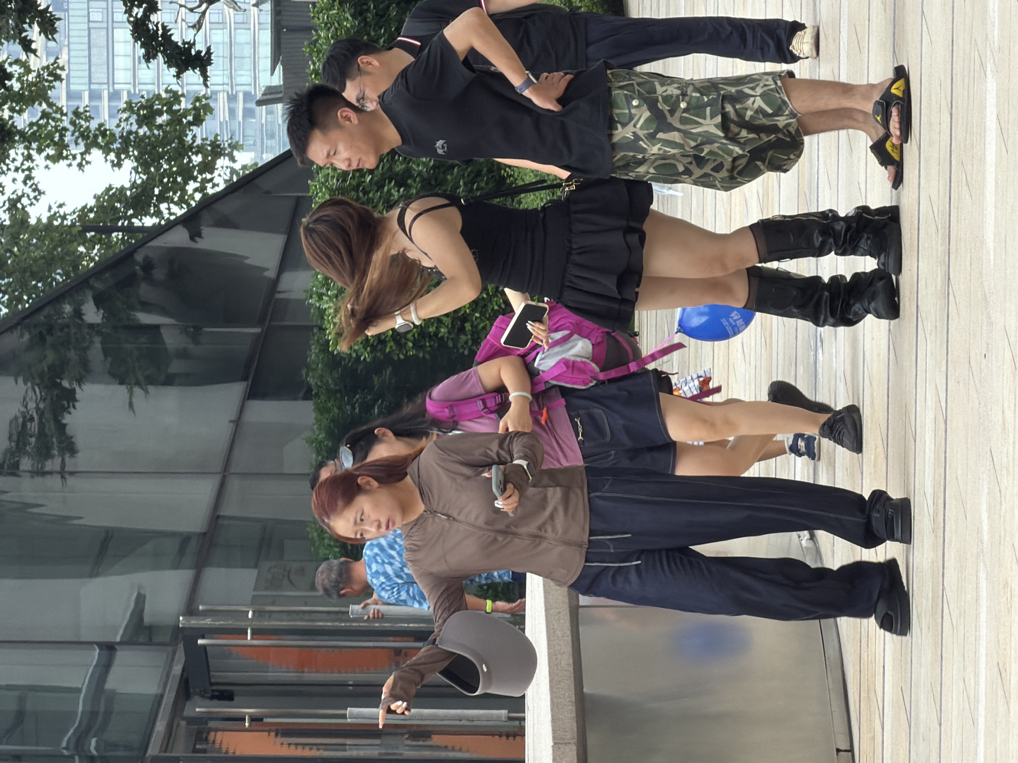 In Yuzhong District, China, a group of people are walking on a tiled sidewalk in front of a modern building with a reflective glass facade. A woman in the foreground, with auburn hair pulled back, wears a brown zip-up jacket over a darker shirt and loose-fitting dark blue pants. She is holding a gray visor in her left hand and pointing forward with her right index finger. Beside her, to the right, a woman with long brown hair, also partly obscured, wears a black tank top and a black ruffled skirt, along with tall black boots. She is holding a smartphone. Behind them, a man in a black t-shirt and camouflage shorts stands with his hands on his hips. Another man in a black t-shirt and dark pants is partially visible behind him. In the background, other individuals and trees are visible, with a tall building in the distance. A blue balloon is on the ground near the woman in the black skirt.