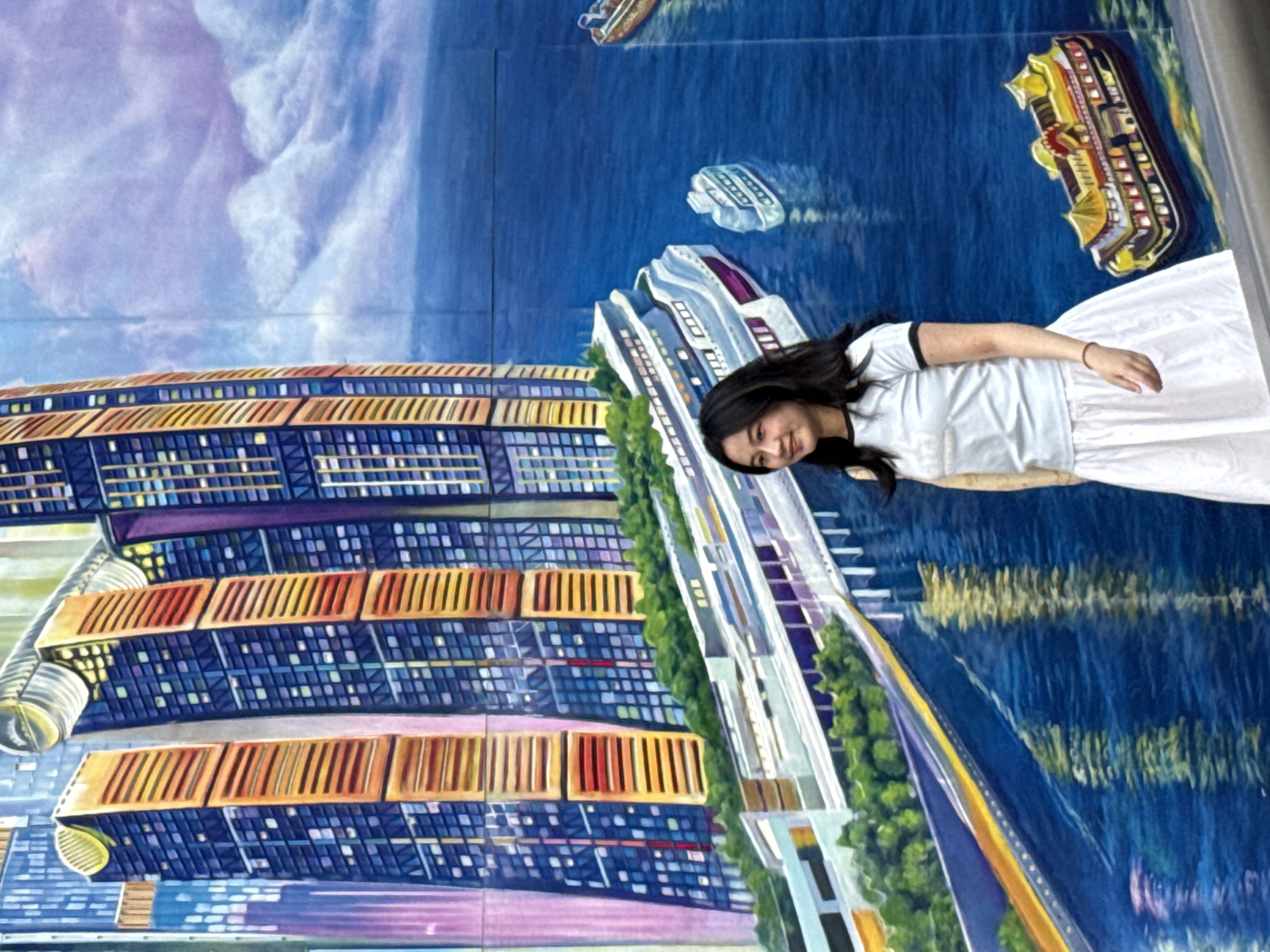 A woman stands in front of a mural, her body angled slightly away from the camera, her face turned towards it, a smile gracing her features. She wears a white T-shirt with dark trim on the sleeves and neck, paired with a white skirt. The mural behind her depicts an urban scene, including buildings, bridges, and boats, suggesting a waterfront city. The painting style is colorful and detailed, evoking the atmosphere of Yuzhong District, China. The environment appears to be outdoors.