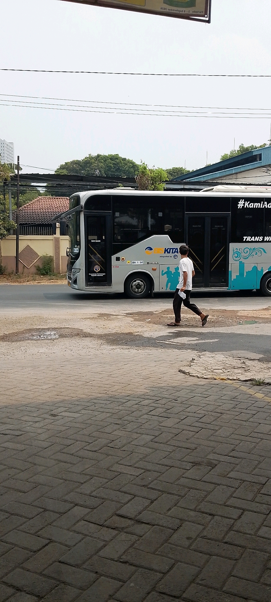 In an urban scene in Bekasi, Indonesia, a man walks across a street in front of a white and blue bus. The bus, which has "KITA" and "#KamiAda" visible on its side, appears to be a public transport vehicle. The man, wearing a white t-shirt and dark pants, carries a white piece of paper. The street surface is a mix of asphalt and paving stones. In the background, there are trees, a building with a tiled roof, and power lines. The sky is overcast.
