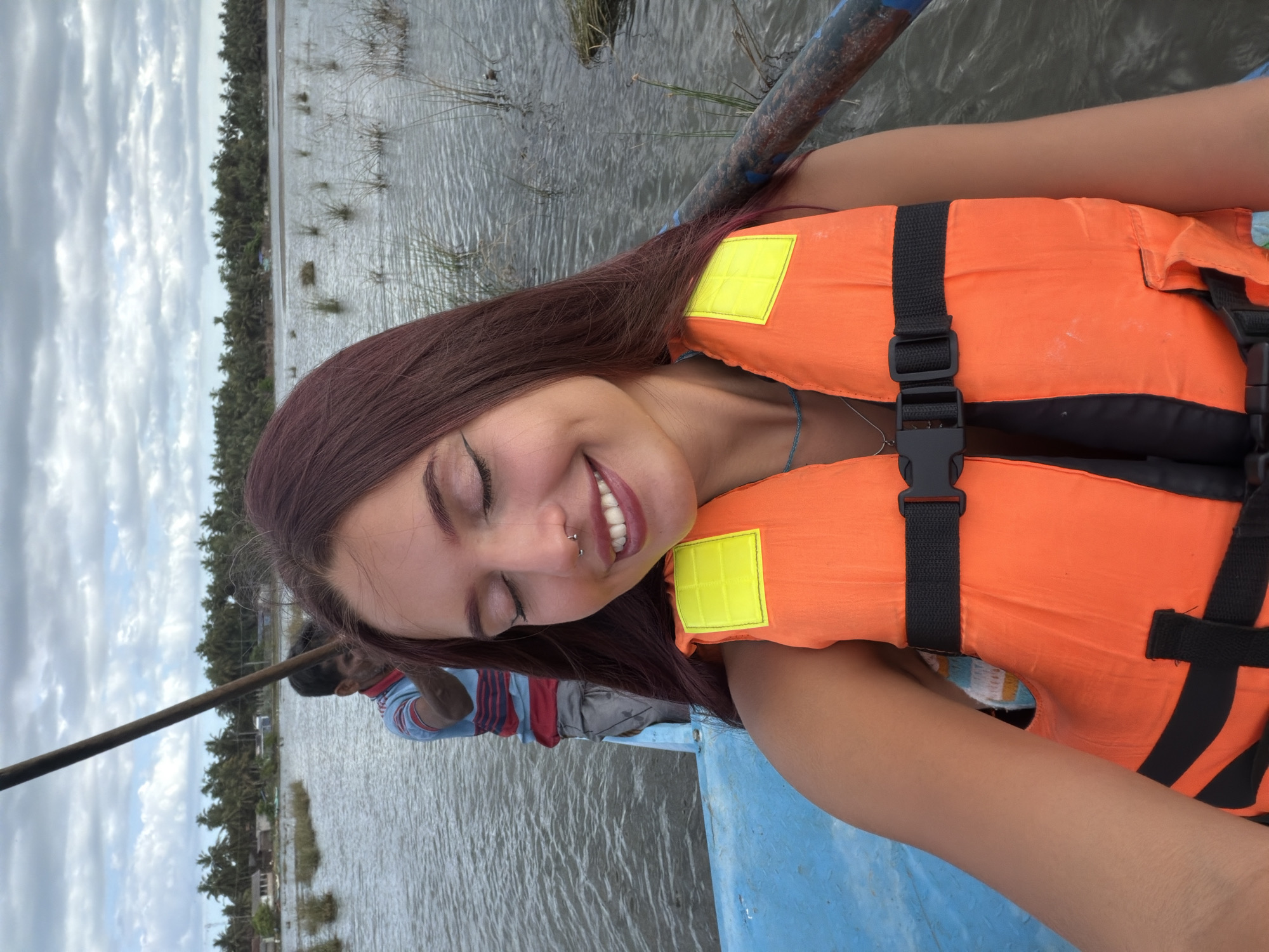 A young woman with long, dark red hair, wearing an orange life vest with yellow reflective patches and a nose ring, smiles with her eyes closed while sitting in a boat. Behind her, a man in a blue and red striped shirt and gray pants is poling the boat with a long wooden pole. The boat is on a body of water, likely a lagoon or river, with reeds and grasses growing in the water. In the background, a line of palm trees and some buildings are visible, suggesting a coastal or tropical location. The sky is cloudy. The scene appears to be in Pottuvil, Sri Lanka, a coastal town known for its lagoons and palm-fringed beaches.