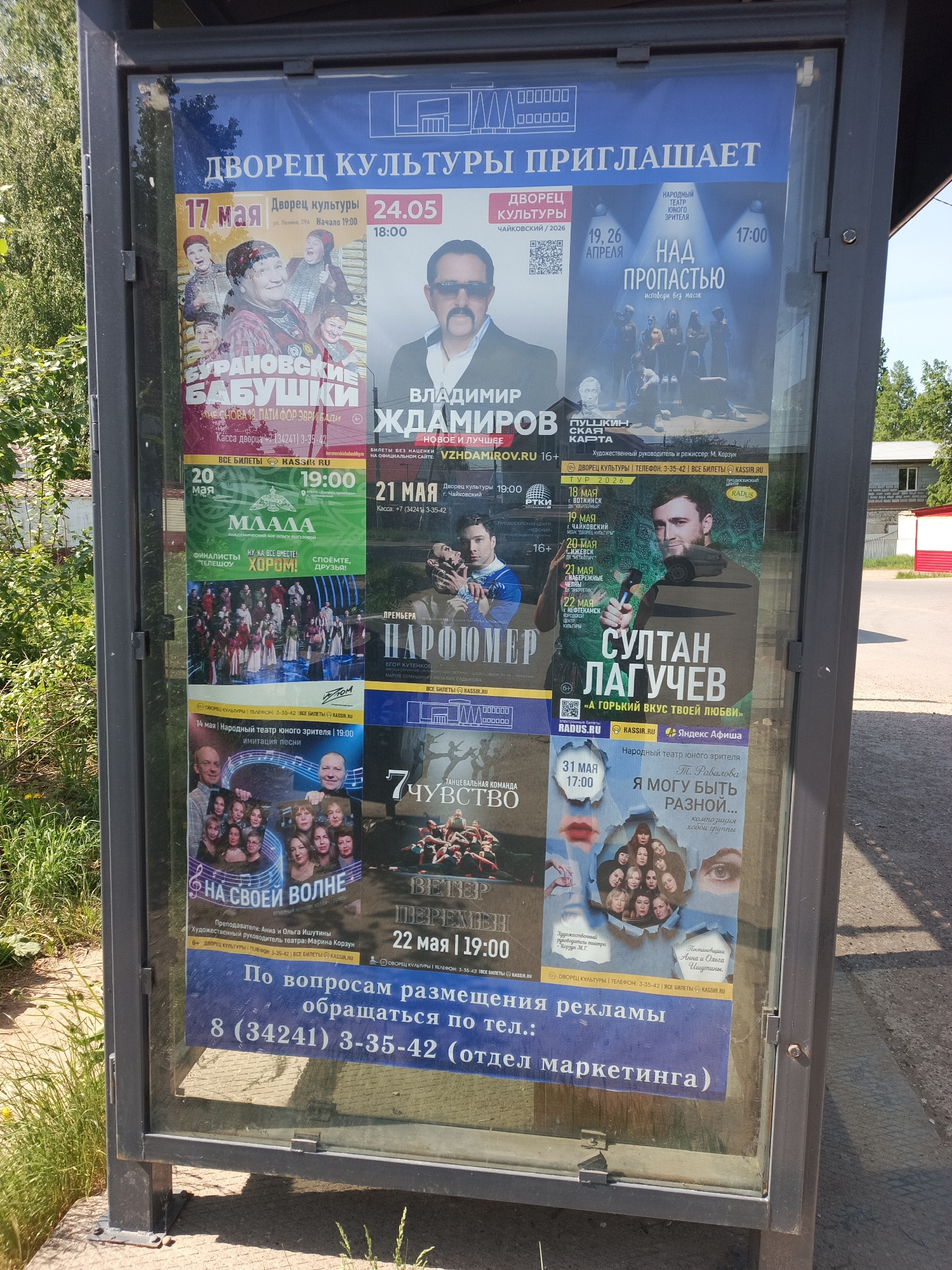 A street-side advertising kiosk displays several promotional posters for events occurring at the “Palace of Culture” (Dvorets Kultury) in the city of Chaikovsky, Russia. The kiosk's location is outdoors. The posters advertise musical performances, theatrical productions, and dance performances. Among the prominent posters are: "Buranovskiye Babushki," "Vladimir Zhdamirov," "Over the Abyss," "Mlada," "Parfumer," "Sultan Laguchev," "On Her Wave," "7th Sense," "Winds of Change," and "I Can Be Different." A phone number is listed at the bottom for advertising inquiries: 8 (34241) 3-35-42 (marketing department). Greenery is visible behind the kiosk. A building with an asphalt road nearby is present as well.