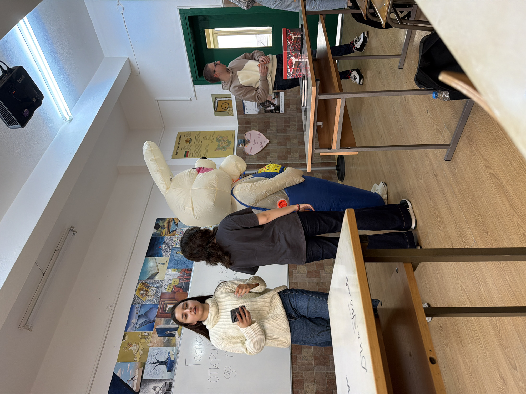 The image shows a classroom in Panagyurishte, Bulgaria. Two women are standing in front of a whiteboard with writing on it. One woman is wearing a white sweater and holding a smartphone. The other woman has dark hair and wears a grey shirt and black pants. Behind them is a large inflatable bunny figure, with a person apparently inside. In the background are students sitting at desks, and a man standing near a door. There is a projector mounted on the ceiling, fluorescent lights, and posters on the walls, including one with a map of Bulgaria. The classroom setting suggests an educational institution.