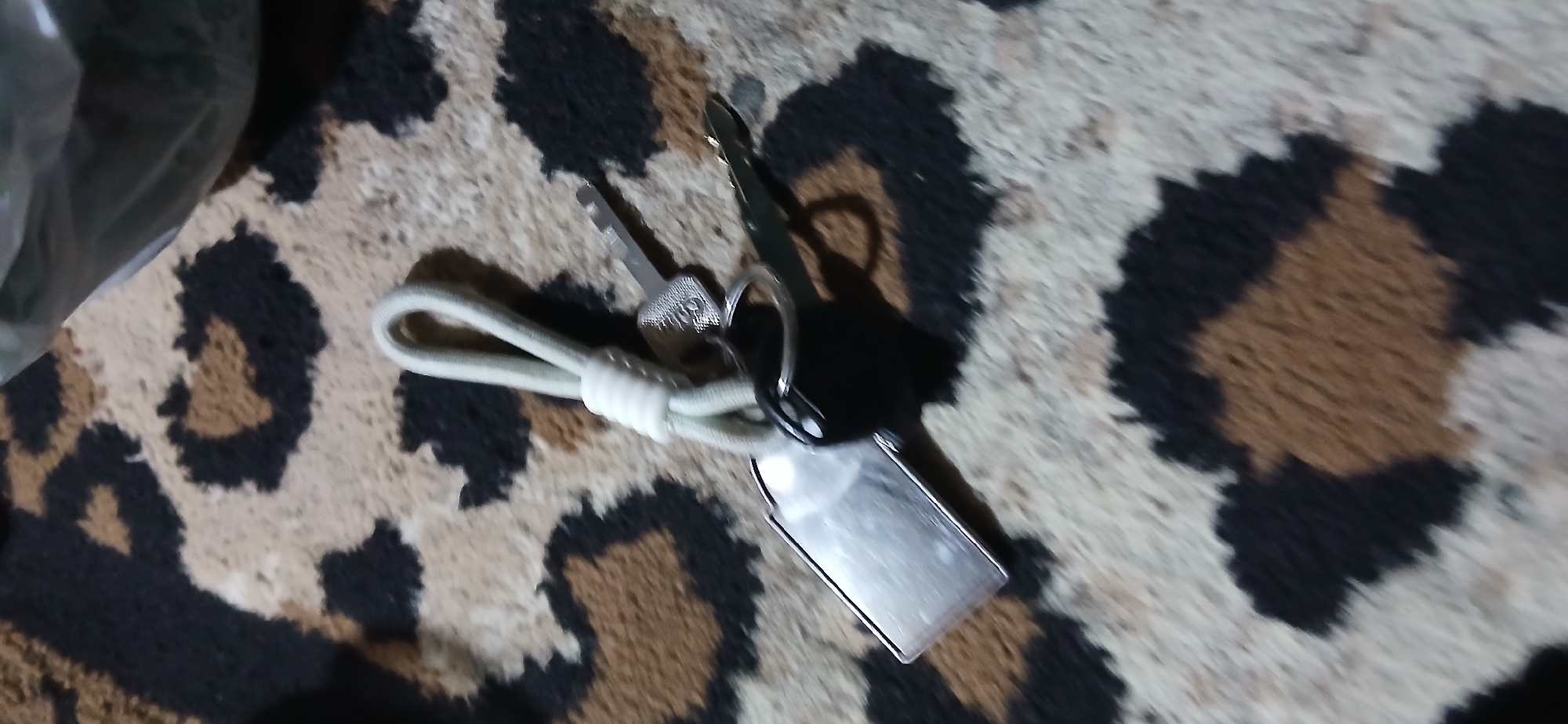 Two car keys and a rectangular metal fob are attached to a light-colored rope keychain. The keys and fob are lying on a carpet with a leopard print pattern. The carpet has patches of black, tan, and cream. The image is taken from a high angle.