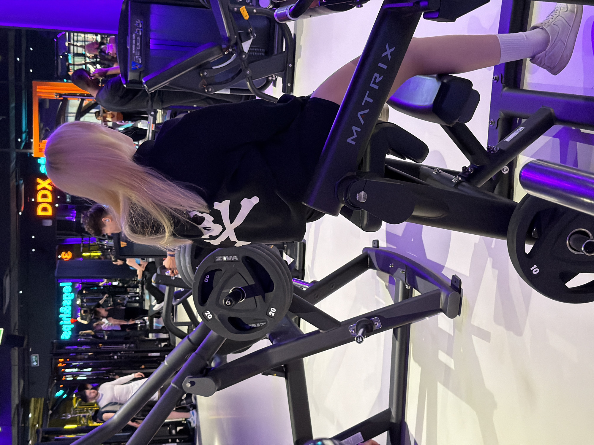 The image depicts a person with blonde hair seated on a Matrix leg extension machine in a gym. The machine has Ziva-branded weight plates on either side. Other gym equipment and people are visible in the background, and a sign with what appears to be neon lighting reads "lɛzgiɛziq". Based on the sign, the location is Saint Petersburg, Russia.