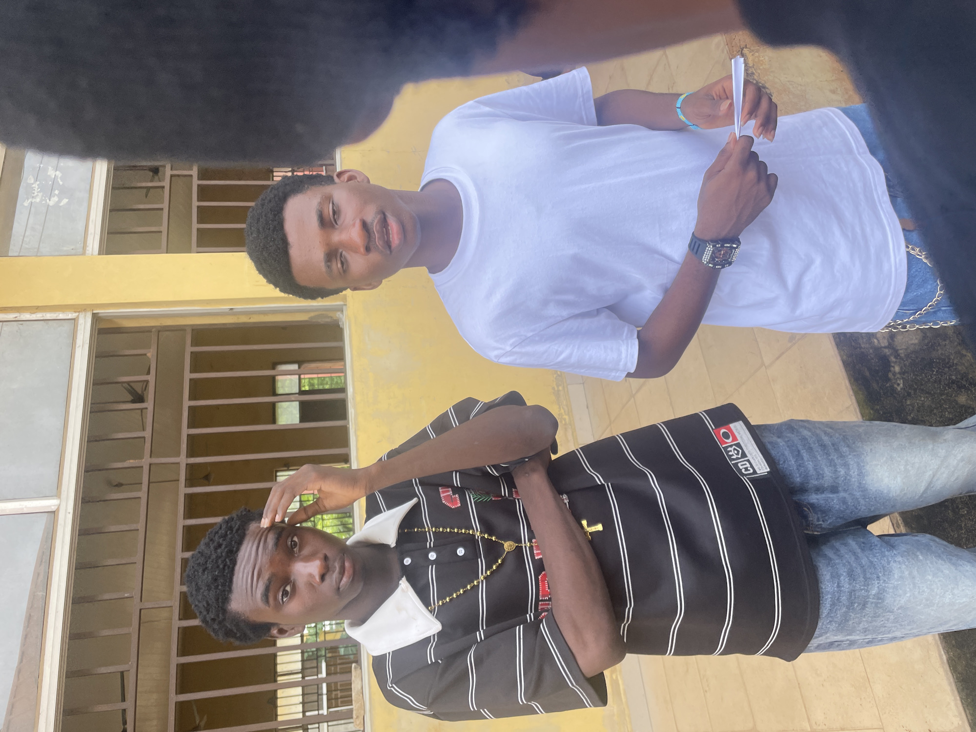 The image depicts two young men standing outside a building with yellow walls and barred windows. The man on the left is wearing a black and white striped polo shirt with a white collar and a gold chain with a cross pendant. He has his arms crossed and his left hand touching his forehead. The man on the right is wearing a white t-shirt and blue jeans. He is holding a small folded piece of paper in his hands. He is also wearing a watch on his left wrist and a blue beaded bracelet on his right wrist. The background shows a walkway or courtyard with a tiled floor. The building's architecture suggests it could be a school or a public institution. The overall setting is likely a warm, outdoor environment. The presence of the building's characteristics and the people's attire suggests a location within the city of Ihiagwa, Nigeria.