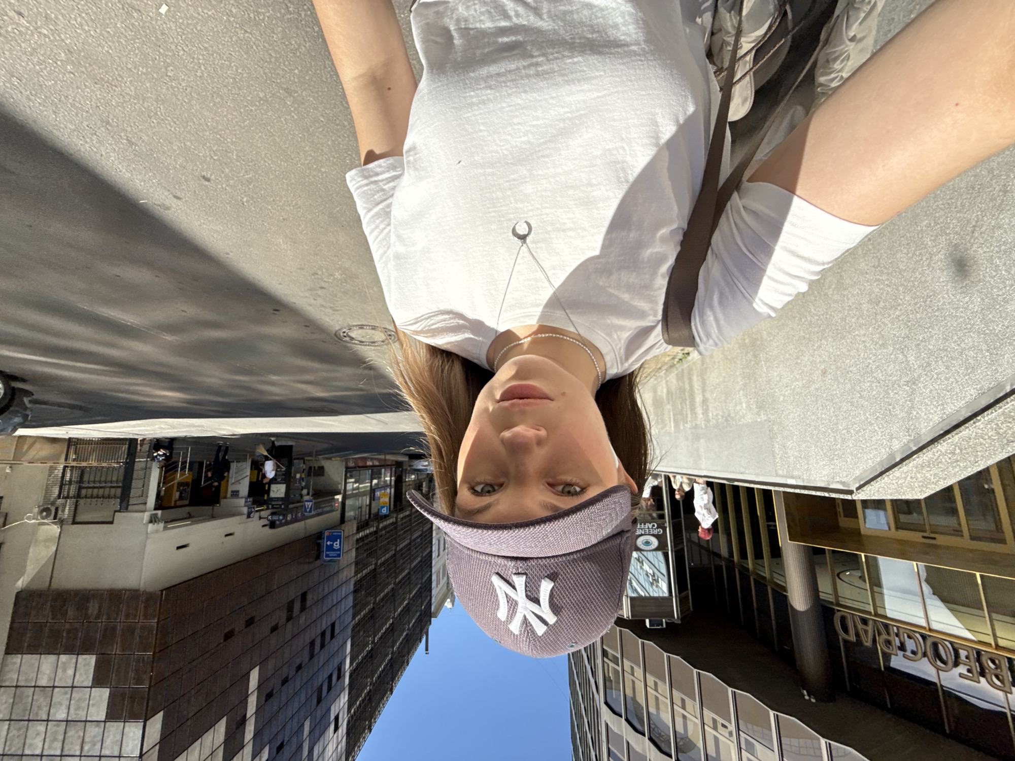 A downward-facing portrait shows a person wearing a gray baseball cap with the New York Yankees logo and a white t-shirt with a silver necklace. Buildings and street scenes are visible in the background, rotated due to the portrait's orientation. A tall building with a grid-like façade is visible on the left, while a sign bearing the word "BEOGRAD" can be seen reflected on a building to the right. Additional buildings and city infrastructure are also visible in the background. The photo was taken on a sunny day. The location is in the city of Vracar Urban Municipality, Serbia.