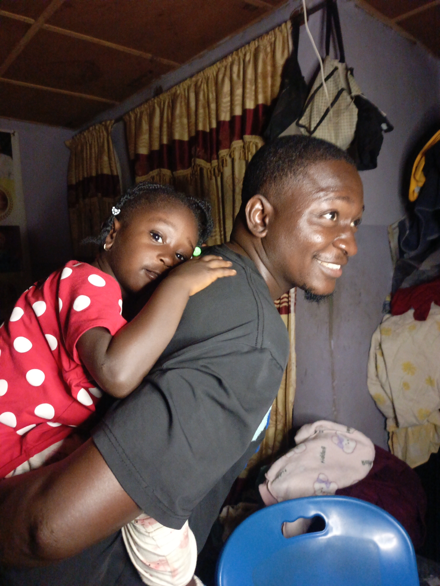 A medium shot captures a Black man and a young Black girl in a dimly lit indoor space. The man is positioned in the foreground, with the girl on his back, resting her head on his shoulder. Both are looking away from the camera. The man wears a black t-shirt, and the girl is dressed in a red t-shirt with white polka dots. The background reveals a wall with curtains and some hanging bags. A blue chair is partially visible in the lower right corner. The environment suggests a casual, domestic setting.