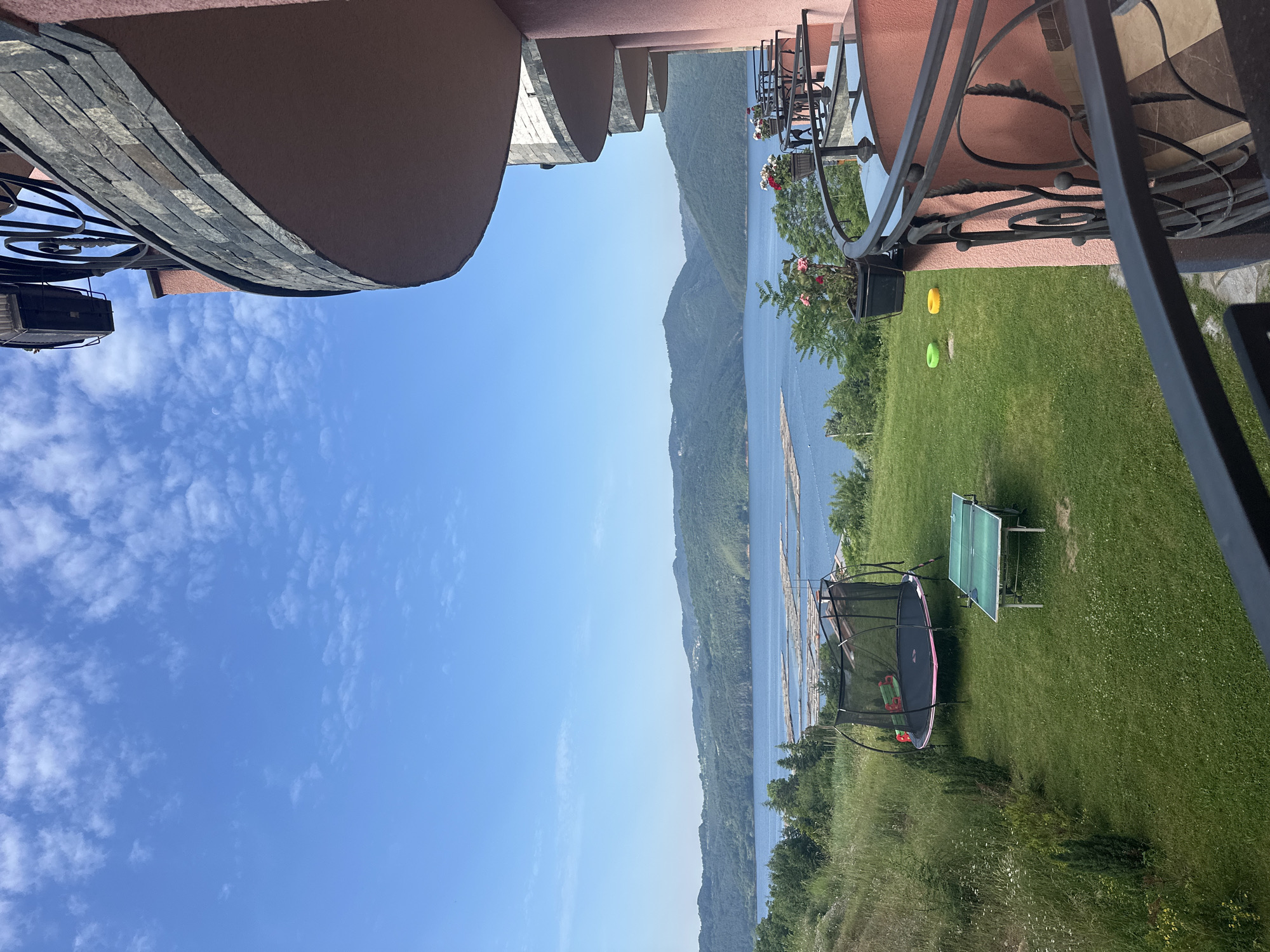 The image depicts a panoramic outdoor scene captured from a multi-level building balcony overlooking a large body of water and forested mountains in Glavatartsi, Bulgaria, under a clear blue sky with scattered cumulus clouds.
In the immediate foreground, a dark metal balustrade with ornate curved patterns frames the lower edge of the view. To the right, a curved building structure with a salmon-pink stucco finish and sections of gray stone cladding is visible. Above the top right balcony, a black light fixture is mounted. Several potted plants with red and white flowers are placed along the balcony railings.
Below the balcony, a well-maintained green lawn occupies the midground. On the lawn, a black-netted trampoline with a pink edge is positioned to the left. To its right, a green foldable ping-pong table stands. Further to the right and closer to the balcony, two colorful plastic rings, one green and one yellow, lie on the grass. The lawn transitions into a patch of wilder, taller grass and small trees on the left slope.
The background features a vast, calm blue lake or reservoir. Numerous light-colored, elongated objects, resembling logs or rafts, are floating on the surface of the water in organized clusters. Beyond the lake, a series of densely forested green mountains rise under the bright daylight sky. A faint, small crescent moon is visible in the upper part of the sky, above the building structure. No individuals are present in the frame.