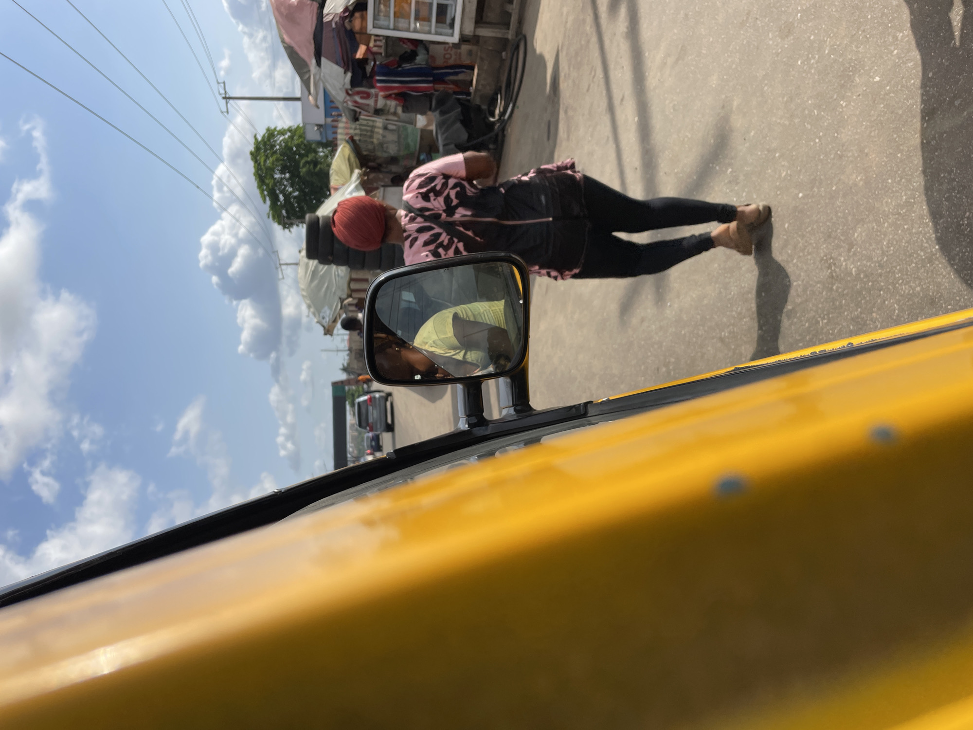 A street scene in Ikeja, Nigeria, as viewed from inside a yellow vehicle. A woman wearing a maroon head covering, a pink and black jacket, and black leggings is standing on the street. Her reflection is visible in the vehicle's side mirror. The street is lined with shops and stalls. The sky is blue with scattered clouds.