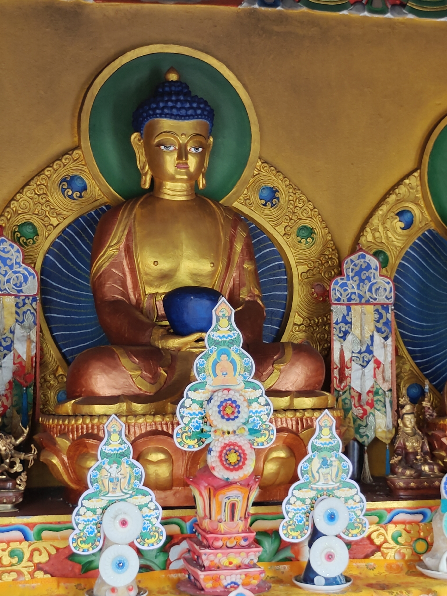 The image depicts a golden statue of Buddha seated in meditation, holding a blue bowl in his lap. The statue is adorned with a green halo and intricate gold detailing on a yellow background. In the foreground, there are smaller, colorful decorative elements, including what appear to be prayer flags and miniature stupas. The setting suggests a Buddhist temple or shrine, likely within Pokhara, Nepal, given the cultural context. The lighting is bright, highlighting the vibrant colors and metallic sheen of the artifacts.