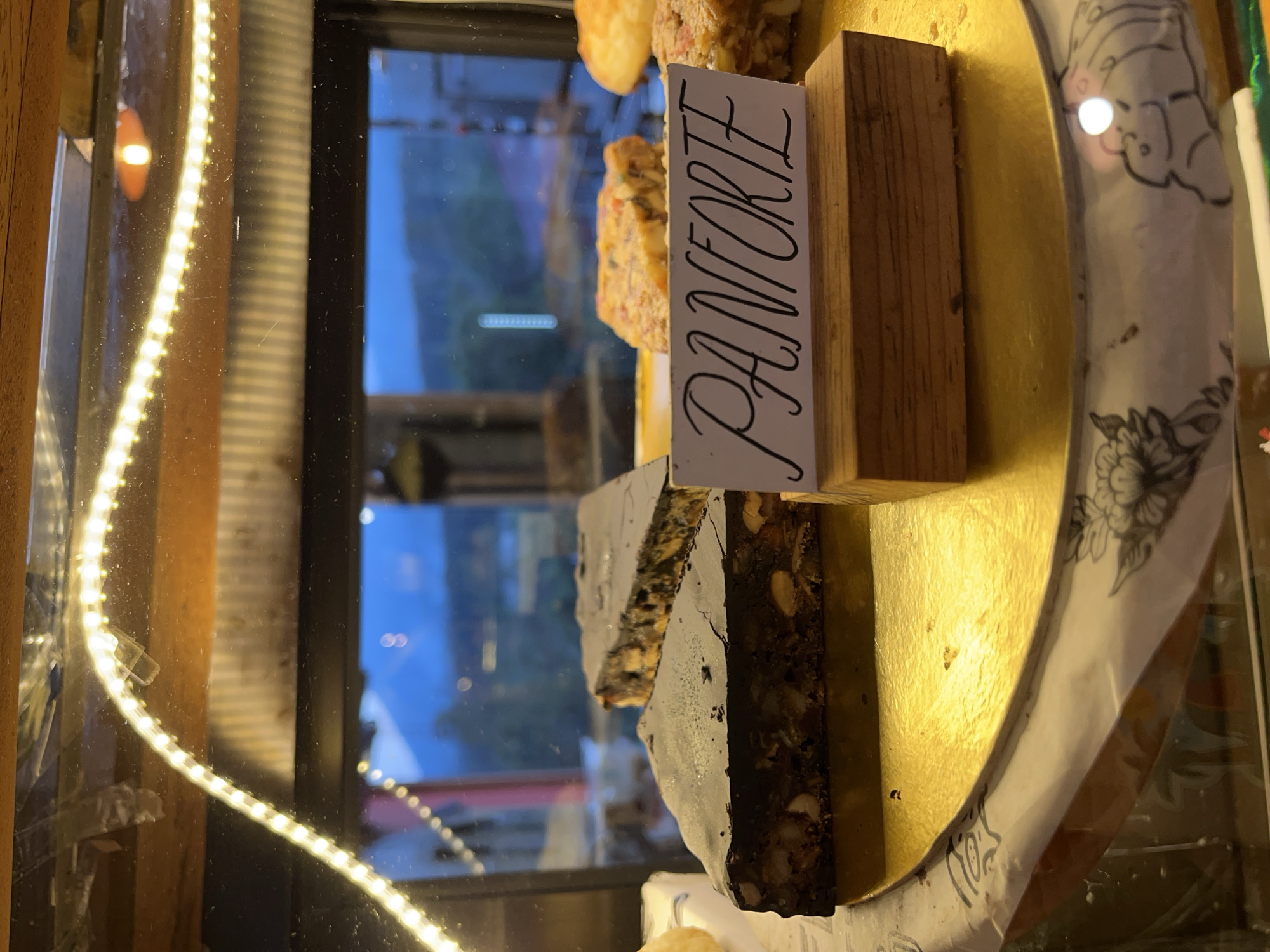 In a glass display case illuminated by an LED strip, a slice of "Panforte" cake sits on a golden platter. The backdrop visible through the glass is the city of Baturiti, Indonesia, appearing slightly blurred.