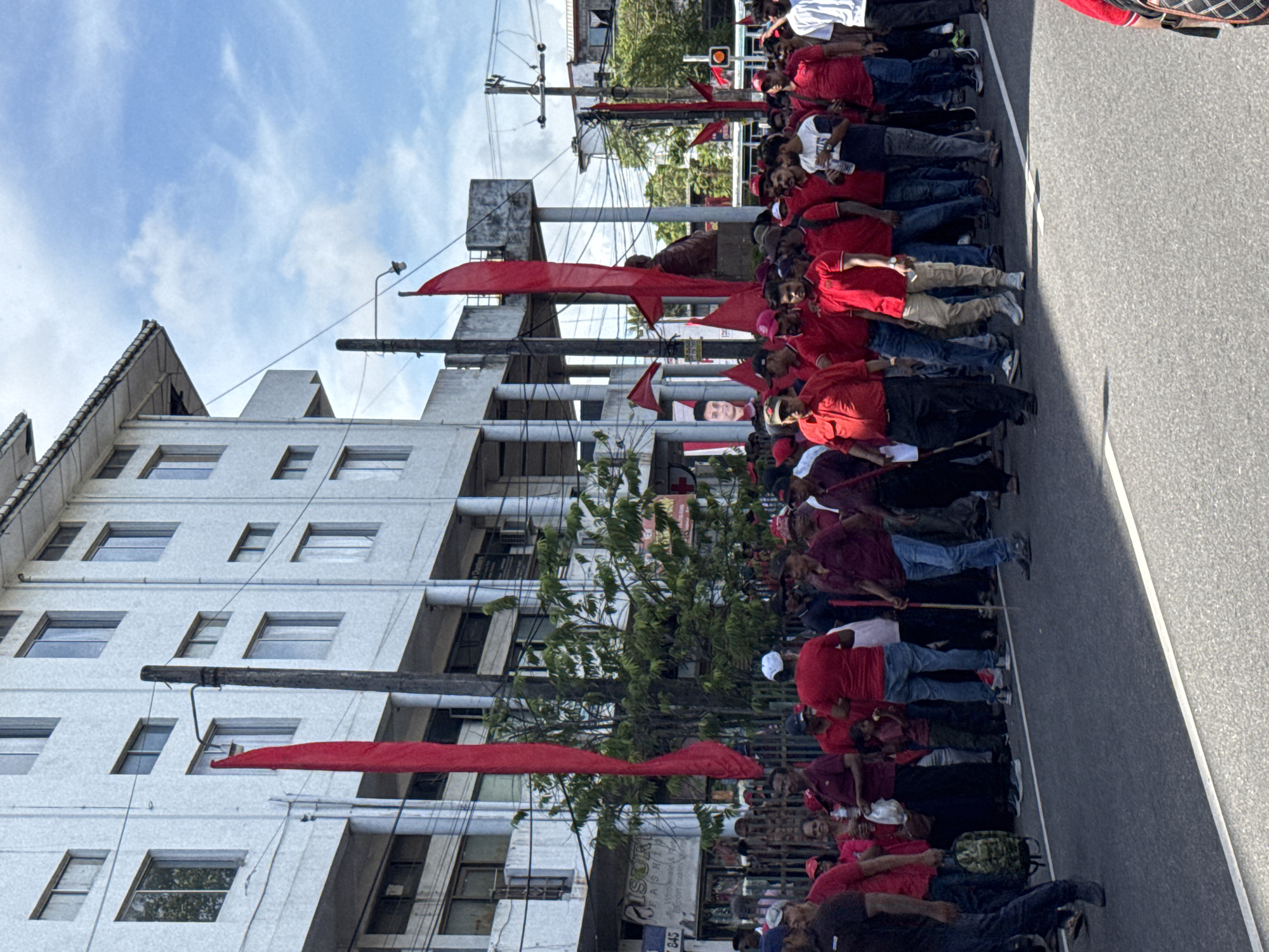 A procession of people, predominantly wearing red shirts and some with red flags, are marching down a street in Delkanda, Sri Lanka. The march is taking place on a paved road next to a multistory building. Overhead power lines are visible, as are trees lining the road. Some individuals are carrying sticks or poles. The building is white with several windows. A billboard is seen above the crowd. A large portrait is visible in the background.