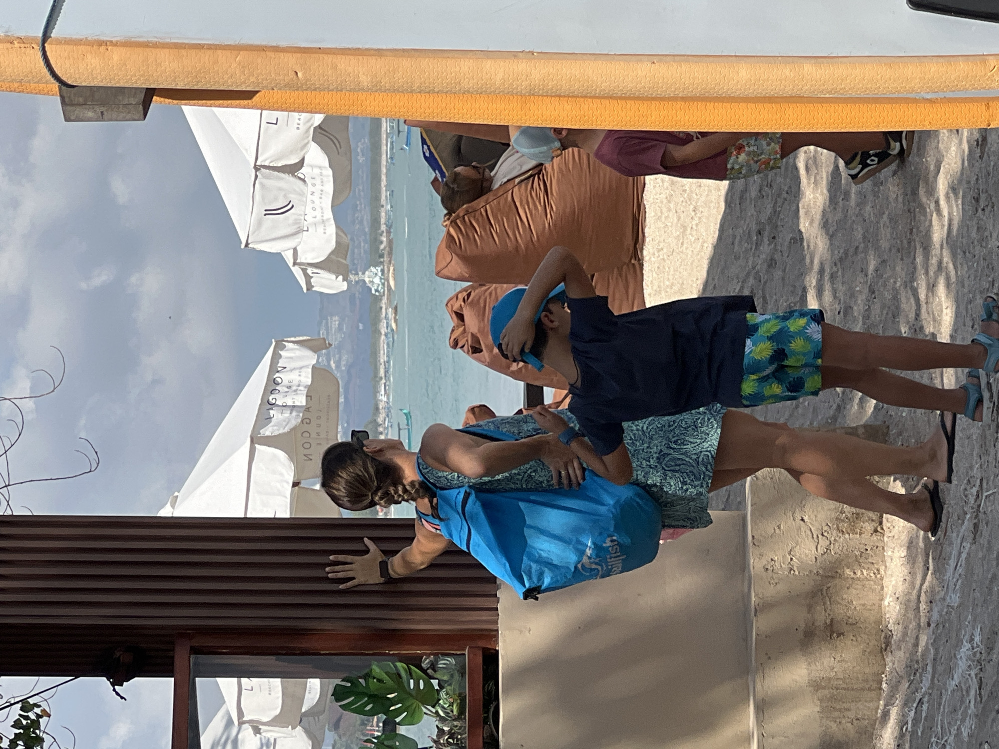 In Kuta, Indonesia, a woman wearing a blue patterned dress and sunglasses stands with her back to the camera, leaning her left hand against a wooden pillar. She has a blue backpack slung over her right shoulder. Beside her, a young boy in a dark blue t-shirt and patterned shorts with a blue cap on his head is looking towards the ocean, his hands behind his head. To the right, another child in a maroon t-shirt and floral shorts is partially visible. In the background, white umbrellas with "Lagoon Lounge" printed on them shade beanbag chairs on a sandy beach, and the turquoise ocean stretches out to the horizon, dotted with boats under a cloudy sky.