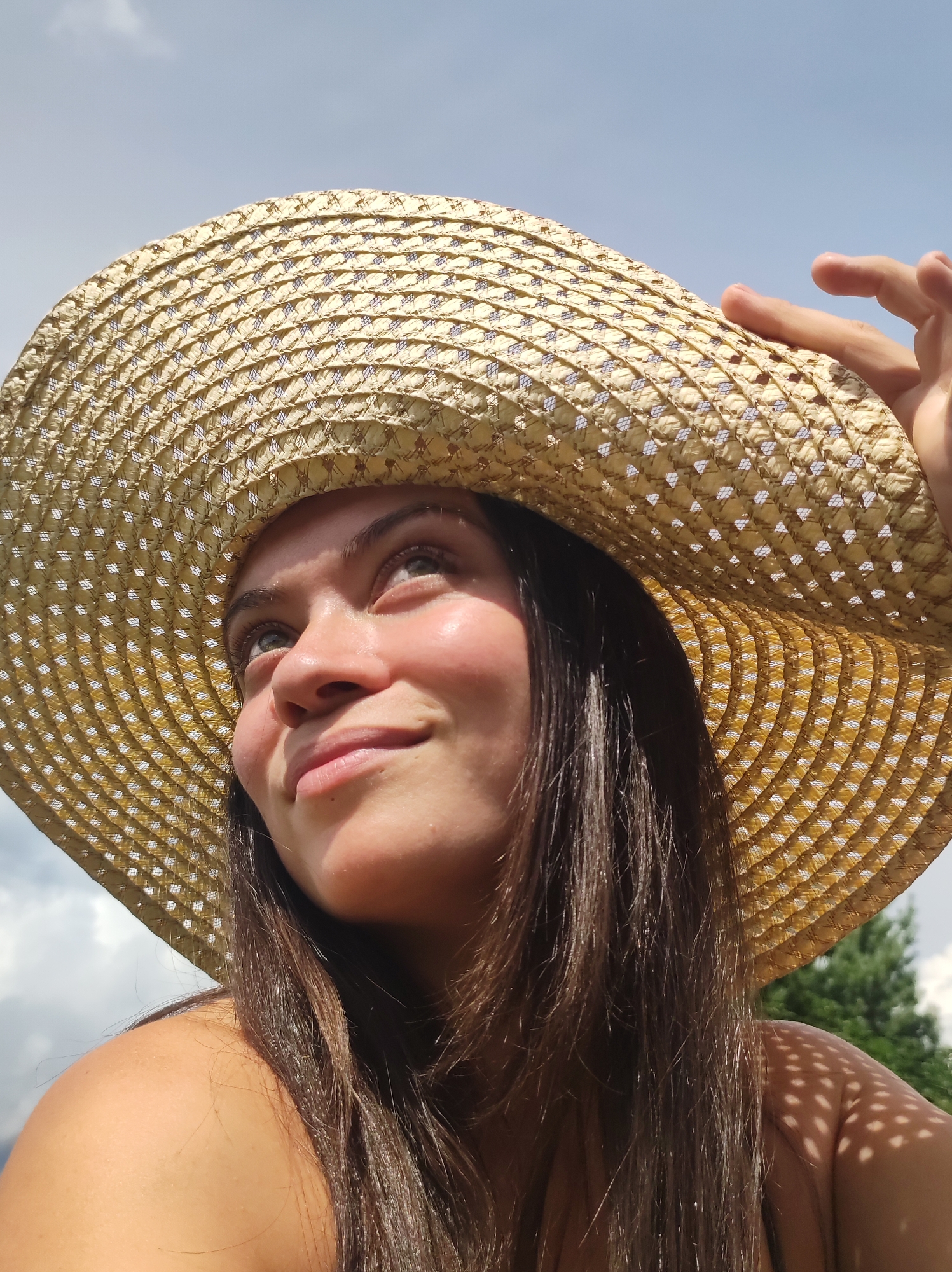 A woman wearing a wide-brimmed straw hat is captured in a close-up, low-angle shot. She is looking upwards and to her left, with a slight smile on her face. Sunlight illuminates her features, creating highlights on her skin and the hat. Her dark hair spills over her shoulders. The background consists of a partly cloudy sky and some greenery, suggesting an outdoor setting. The image does not provide any information about the city of Фоки, Russia.