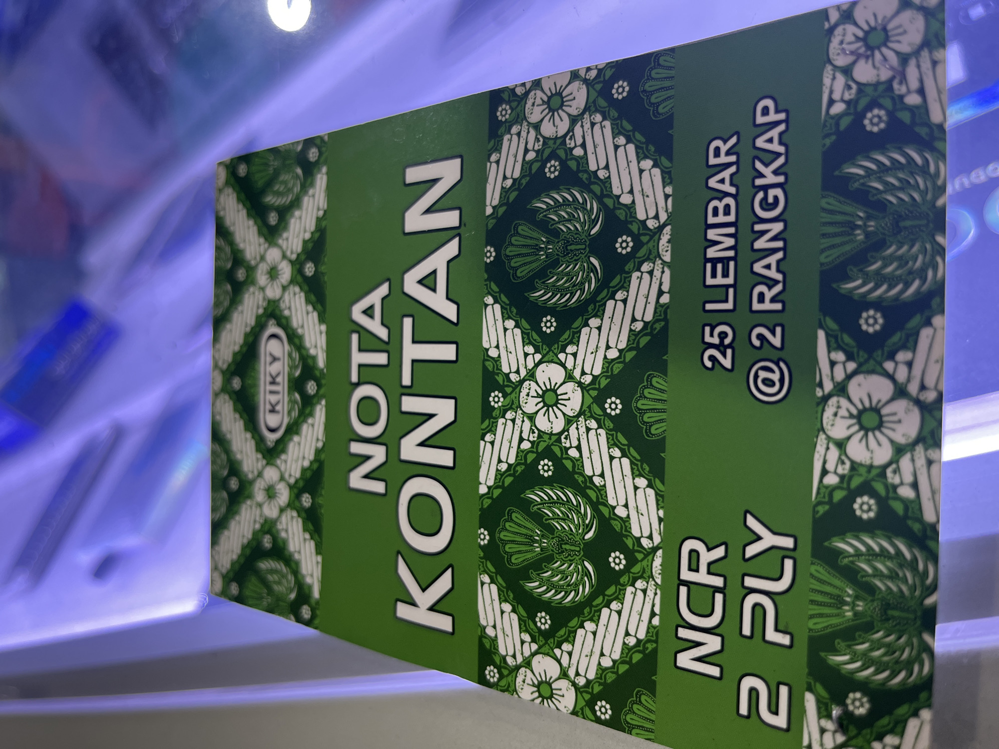 The image shows a close-up, angled view of a green and white patterned notepad. The notepad features a batik-inspired design with floral motifs and what appears to be a crest or emblem in the center of one section. Text on the notepad is in white with a subtle outline. The visible text reads "KIKY" in a circular logo, "NOTA KONTAN", "NCR 2 PLY", and "25 LEMBAR @ 2 RANGKAP". The notepad is likely a carbonless copy form. The background is out of focus and appears to be an indoor setting with bright, possibly fluorescent or LED lighting casting a purple and blue hue. The surface the notepad is resting on is reflective and shows some indistinct shapes and colors in the background. There are no people or specific city landmarks visible.