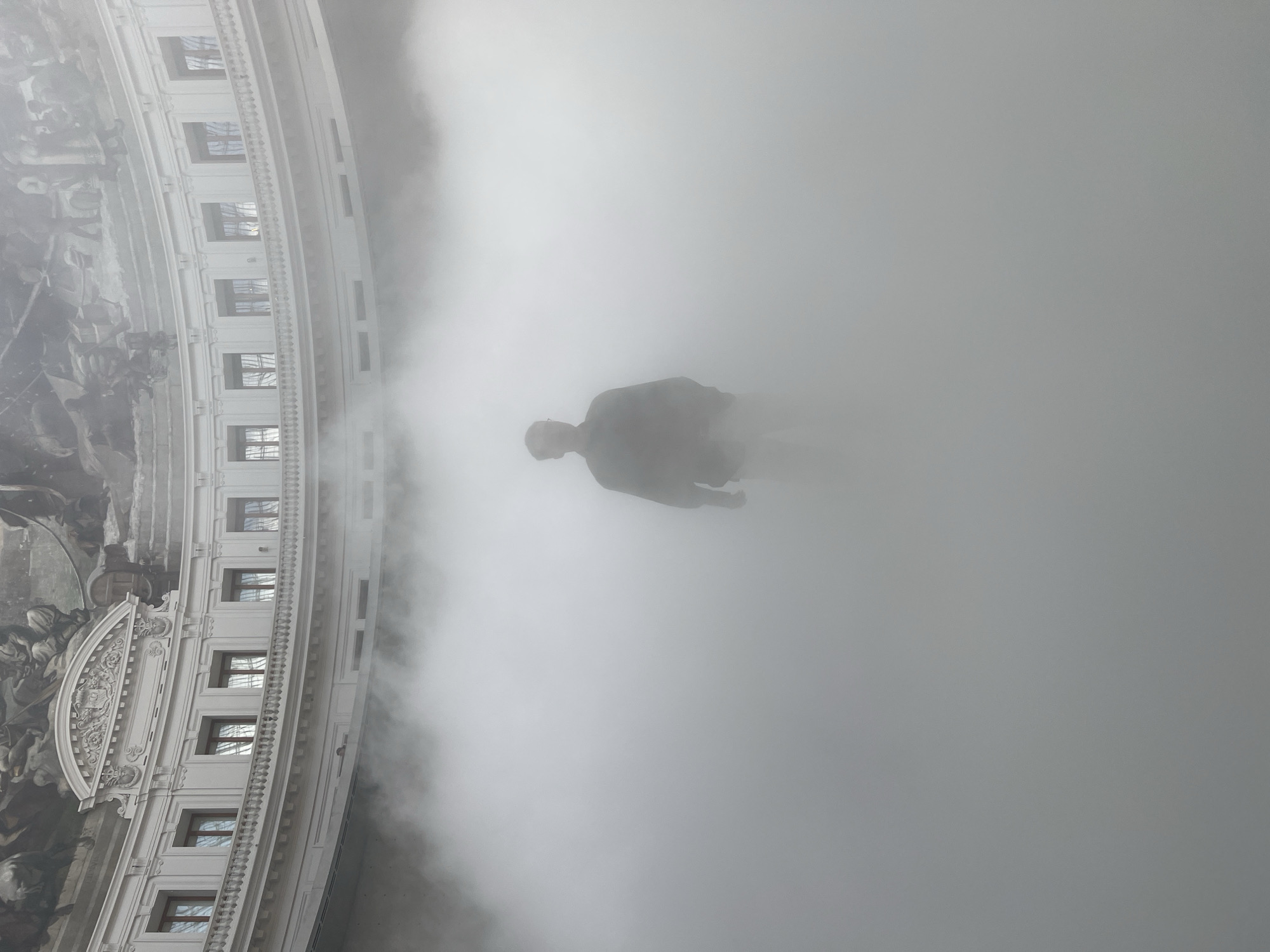 A single individual stands in the center of a circular, white-walled atrium filled with dense white mist. The person is silhouetted against the bright mist, wearing a dark jacket and lighter pants, with hands in pockets. Above the mist, the upper level of the atrium is visible, featuring a curved facade with regularly spaced windows and decorative architectural elements, including a mural on the domed ceiling. The mist obscures the lower portion of the atrium and most of the surrounding architecture, creating a sense of depth and mystery. This scene is captured within the Panthéon in Paris, France.