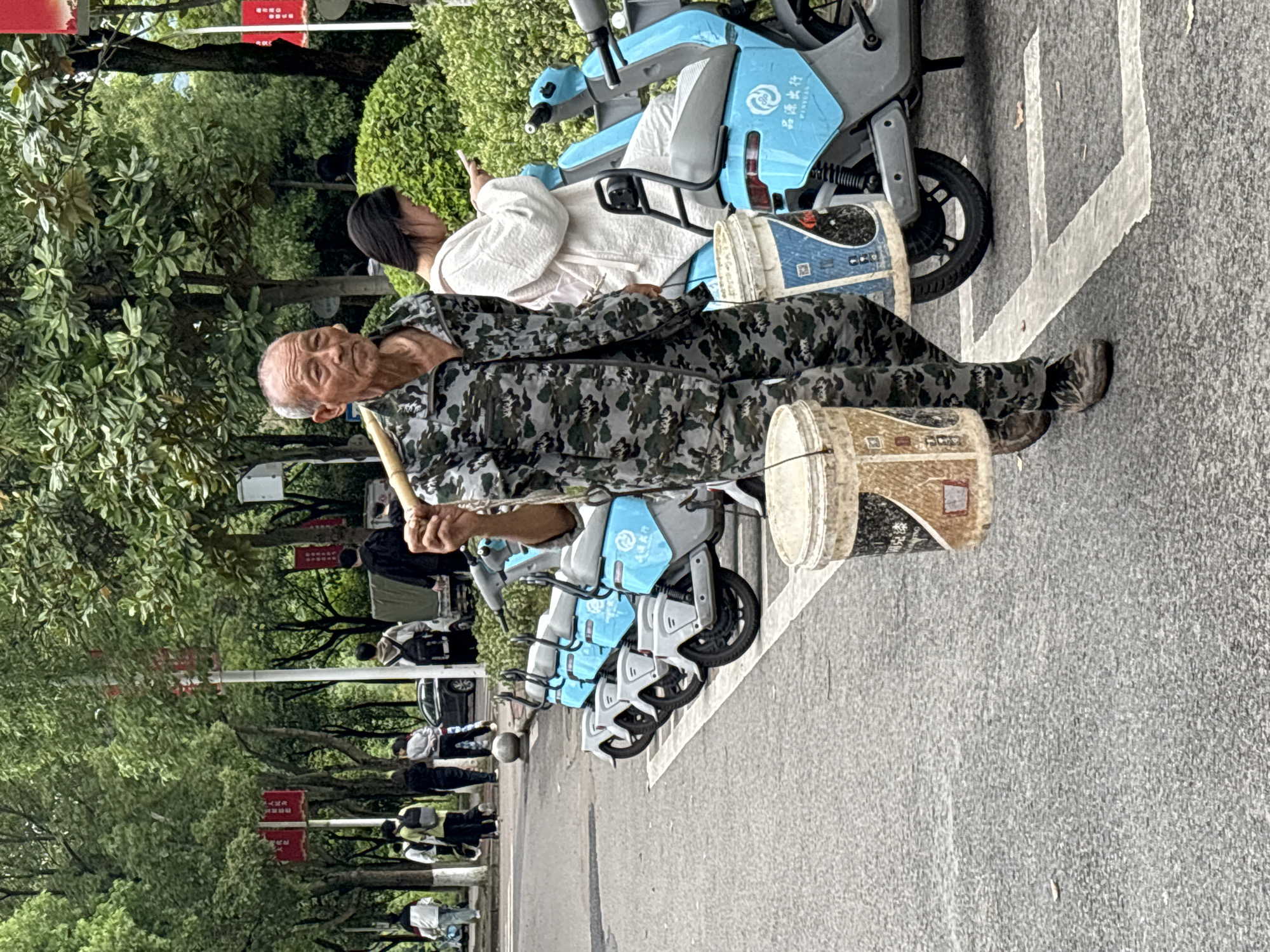 The image, taken in Jiangjin, China, depicts an outdoor scene featuring an elderly man, a young woman, and several shared electric scooters. In the foreground, an elderly man with a weathered face and short hair is captured, wearing a camouflage jacket and trousers. He holds a wooden stick in his right hand, poised as if performing percussion on one of the two light-colored buckets placed beside him. The buckets appear to be made of plastic or similar material, with one displaying some faded text and graphics.
Behind the elderly man, a young woman with dark hair is seated on a light blue and white shared electric scooter, engrossed in her mobile phone. She is wearing a light-colored, possibly white, long-sleeved top. Several identical light blue and white shared scooters are parked in a row behind them. The brand "PINGYUAN 品源出行" is visible on the side of one of the scooters.
The setting appears to be a paved area or sidewalk, with white markings on the ground suggesting a designated parking spot. The background is dominated by lush green trees and bushes, indicating a park-like or well-vegetated urban environment. In the distance, other pedestrians can be seen, and red signs with Chinese characters are visible hanging from posts among the trees.
The lighting suggests it is daytime, possibly overcast, as there are no strong shadows. The elderly man's posture and the buckets imply he is a street performer or busker. He looks towards the viewer with a slight expression, while the young woman is focused on her phone. The scene conveys a lively, everyday atmosphere in a public space.