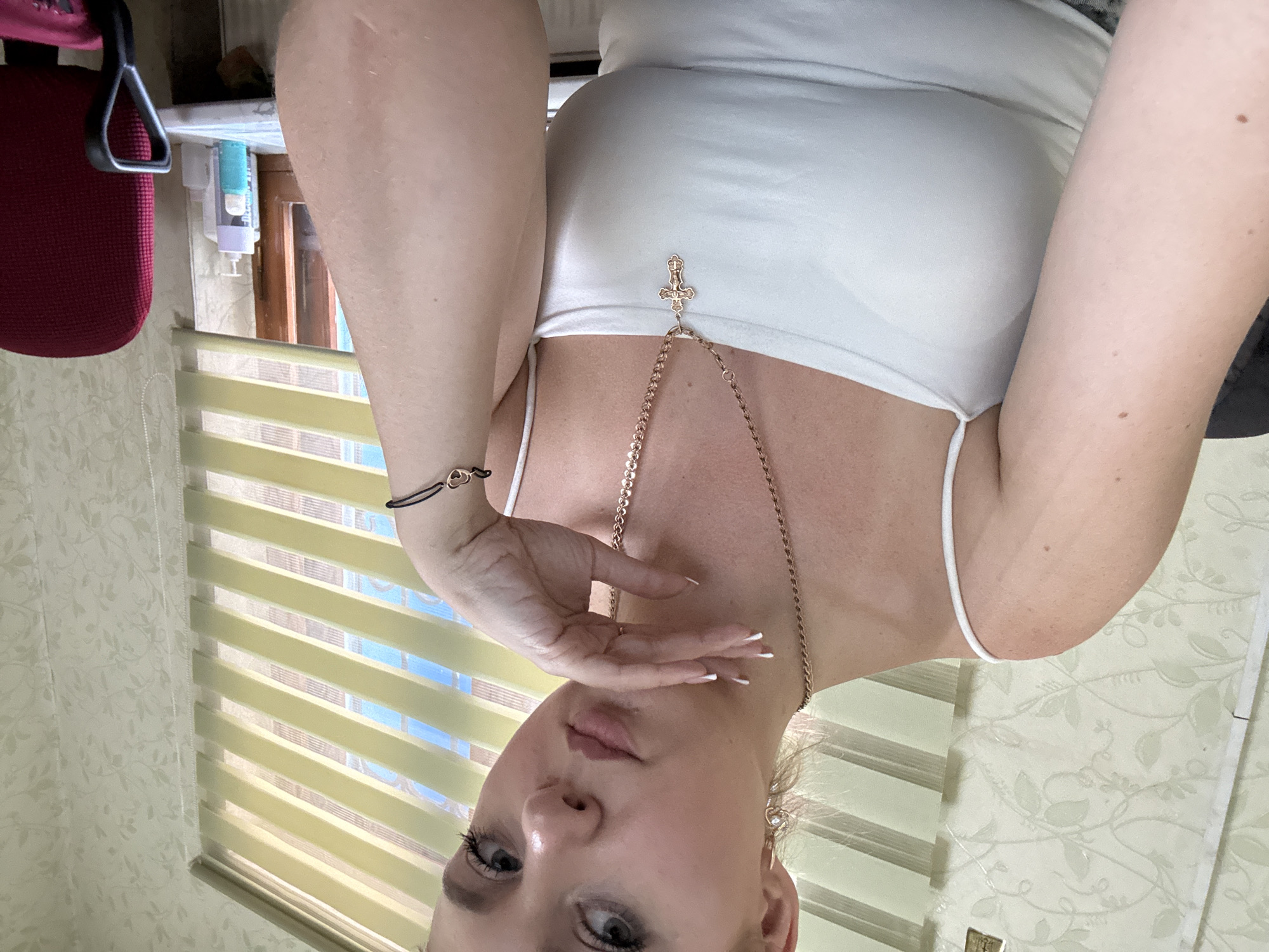 A woman is posing for a selfie, her body is angled towards the camera. She is wearing a white tank top and a rose gold necklace with a cross pendant. She has a black string bracelet on her left wrist with a heart charm. Her hand is raised to her face, with her fingers delicately touching her cheek. The background shows a window with horizontal blinds and a patterned wallpaper. There are some bottles on a shelf behind her. The image is taken indoors, likely during the daytime. The woman's expression is neutral. There is no visible text in the image.