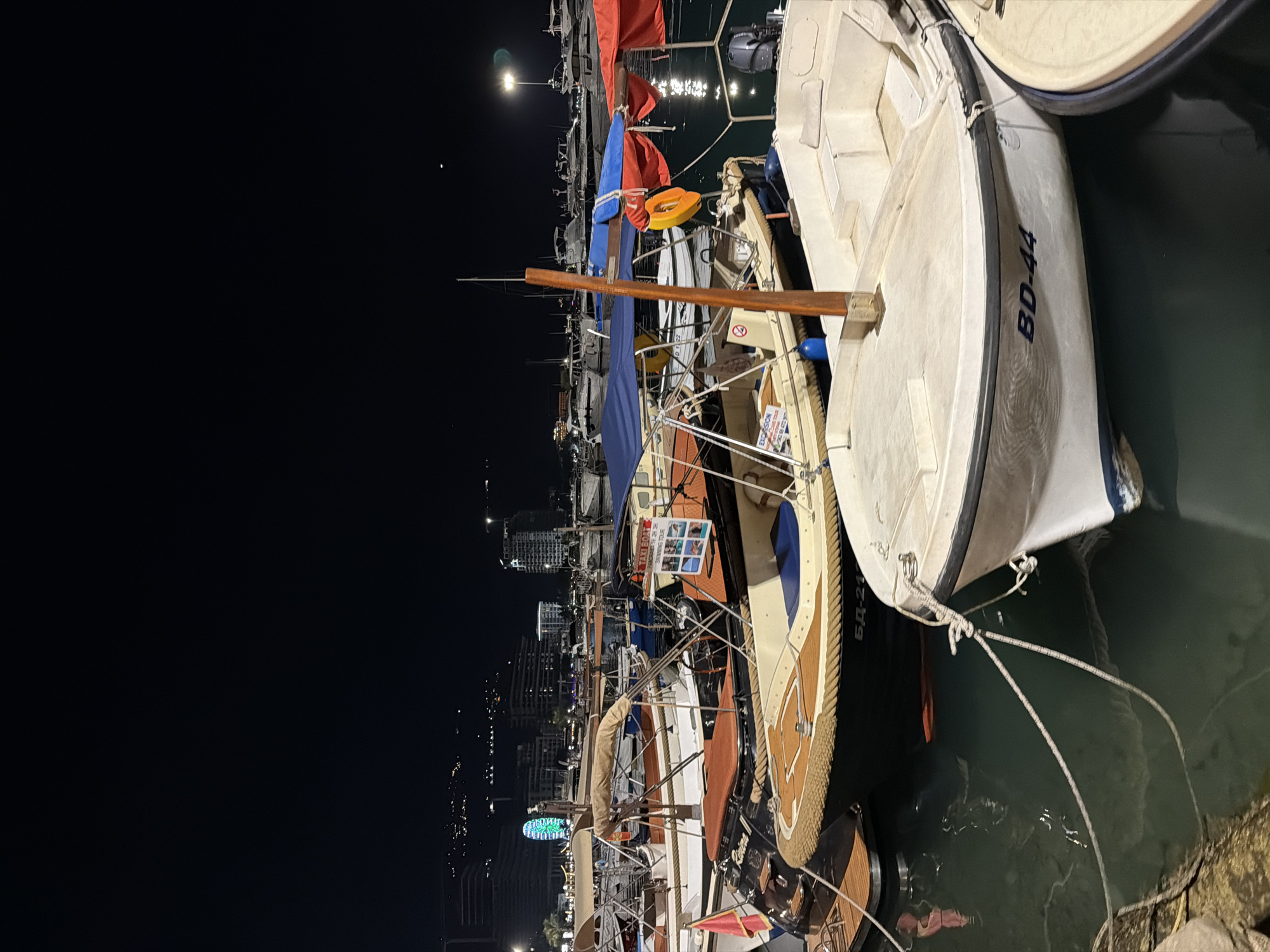 The image depicts a nighttime scene at a harbor in Budva, Montenegro. Several boats are docked, with the foreground dominated by a white sailboat featuring the registration number "BD-44" on its hull. The sailboat has a wooden trim and is tied to the dock with ropes. Various other boats are visible behind it, some with colorful sails and life preservers.
In the background, a cityscape with illuminated buildings and a Ferris wheel can be seen. The water in the harbor reflects the lights from the city and the boats. The sky is dark, suggesting it is nighttime, and a bright light source, possibly a moon or a streetlamp, is visible in the upper left corner.
One of the boats has a sign attached to it with images of people swimming and text that appears to be an advertisement for swimming tours. The overall atmosphere is calm and serene, with the lights creating a warm glow on the water.