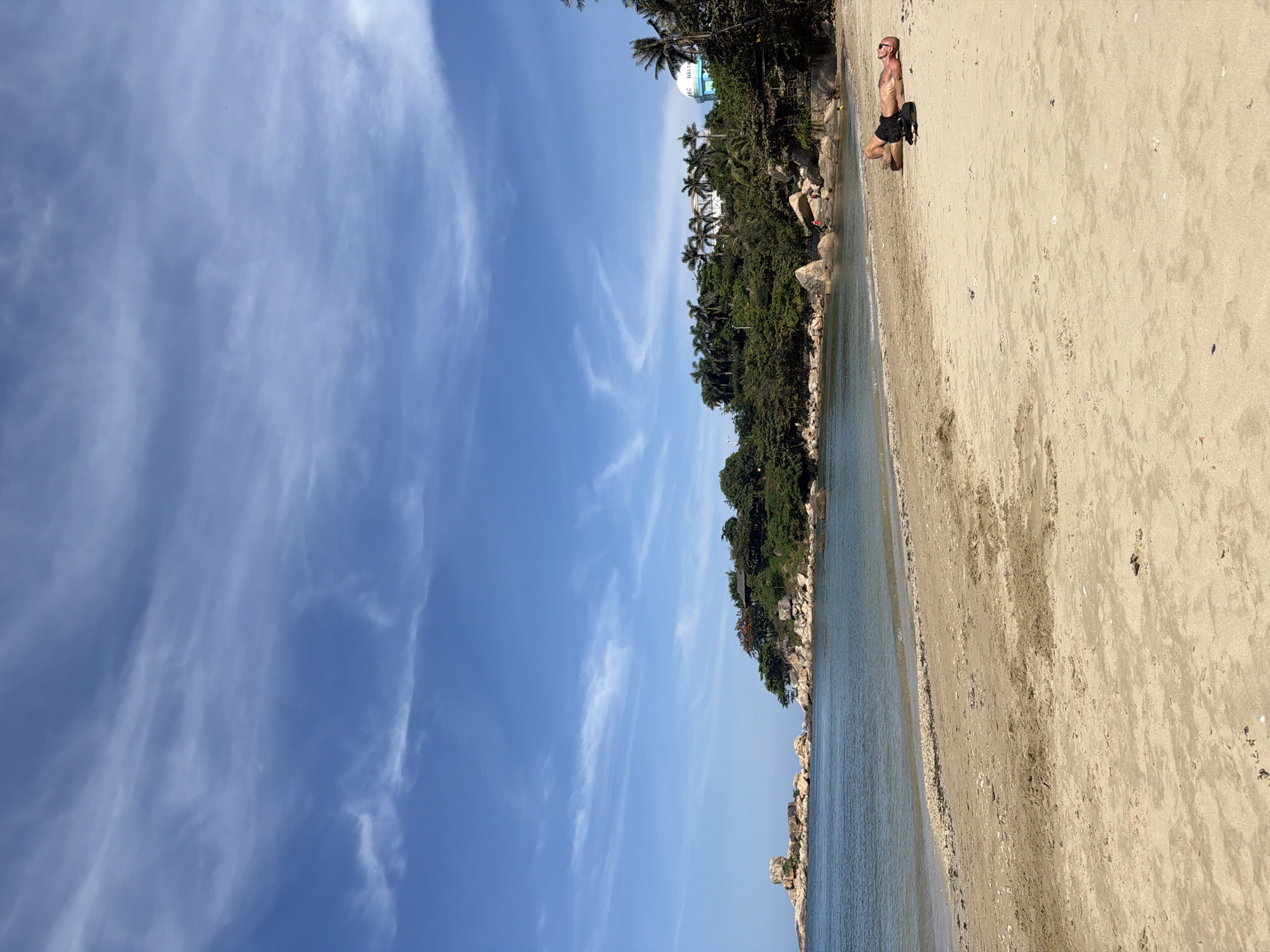 The image shows a beach scene in Bac Nha Trang Ward, Vietnam. A fair-skinned male with a shaved head, wearing sunglasses and dark shorts, is lying on his side on the sand. He appears to have a dark bag or object beside him. The beach is sandy and slightly wet closer to the water. Lush green vegetation, including palm trees, covers the terrain adjacent to the beach. Large rocks are visible along the shoreline. The sky is blue with wispy cirrus clouds. A tall, white, cylindrical structure with "I-SSC UNITED" written on it can be seen among the trees.