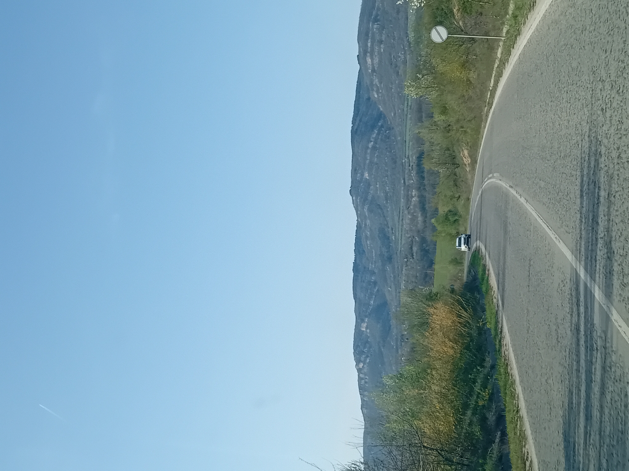 The image depicts a winding road with a white line on its right side, curving towards the right of the frame. On the right side of the road, there is vegetation, including trees and grass. A white vehicle is visible on the road in the distance. Adjacent to the vegetation, there is a mountain range in the background. The sky is blue and mostly clear, occupying a significant portion of the left side of the image. At the upper right, a road sign is visible. The location is Unknown, Unknown.