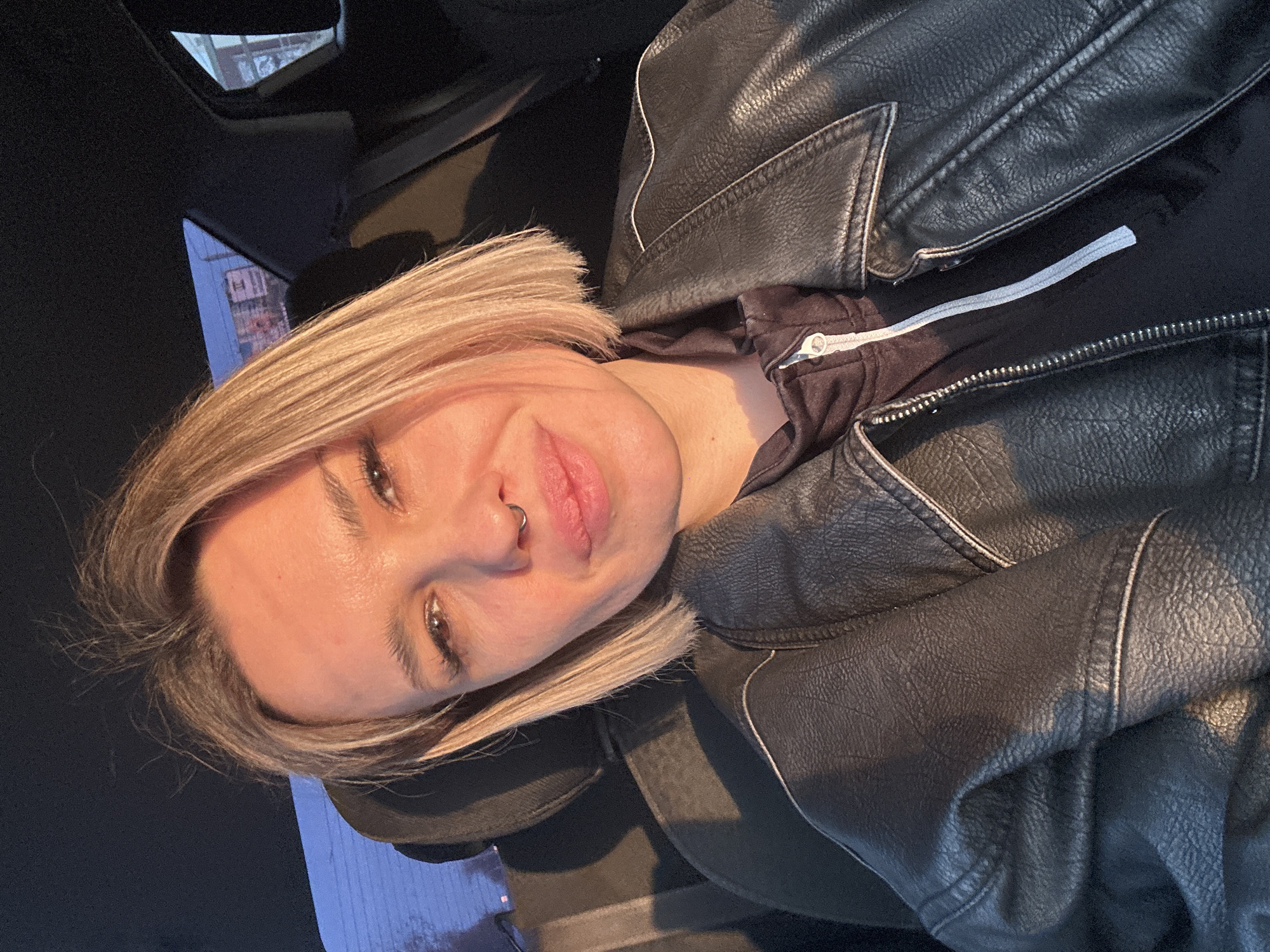 An adult female is depicted seated inside the dark interior of a vehicle, appearing to take a self-portrait. She has shoulder-length, straight blonde hair, brown eyes, and a septum piercing. Her lips are slightly parted, and she looks directly at the camera with a subtle smirk. She is wearing a black faux leather jacket over a dark zip-up hoodie, which features a prominent white zipper pull. Strong, warm, directional lighting, consistent with late afternoon or early morning sunlight, illuminates the left side of her face and body from the viewer's perspective, casting shadows. Through the vehicle's rear windows, an urban environment in Rostov-on-Don, Russia, is visible. The background includes the upper floors of a multi-story building, appearing to be commercial or mixed-use, with some reddish and lighter-toned elements and possible signs. Utility lines or structures are faintly discernible against a twilight sky that transitions from light blue at the horizon to darker blue higher up. Specific street or landmark identification within Rostov-on-Don is not possible from the visible background details.