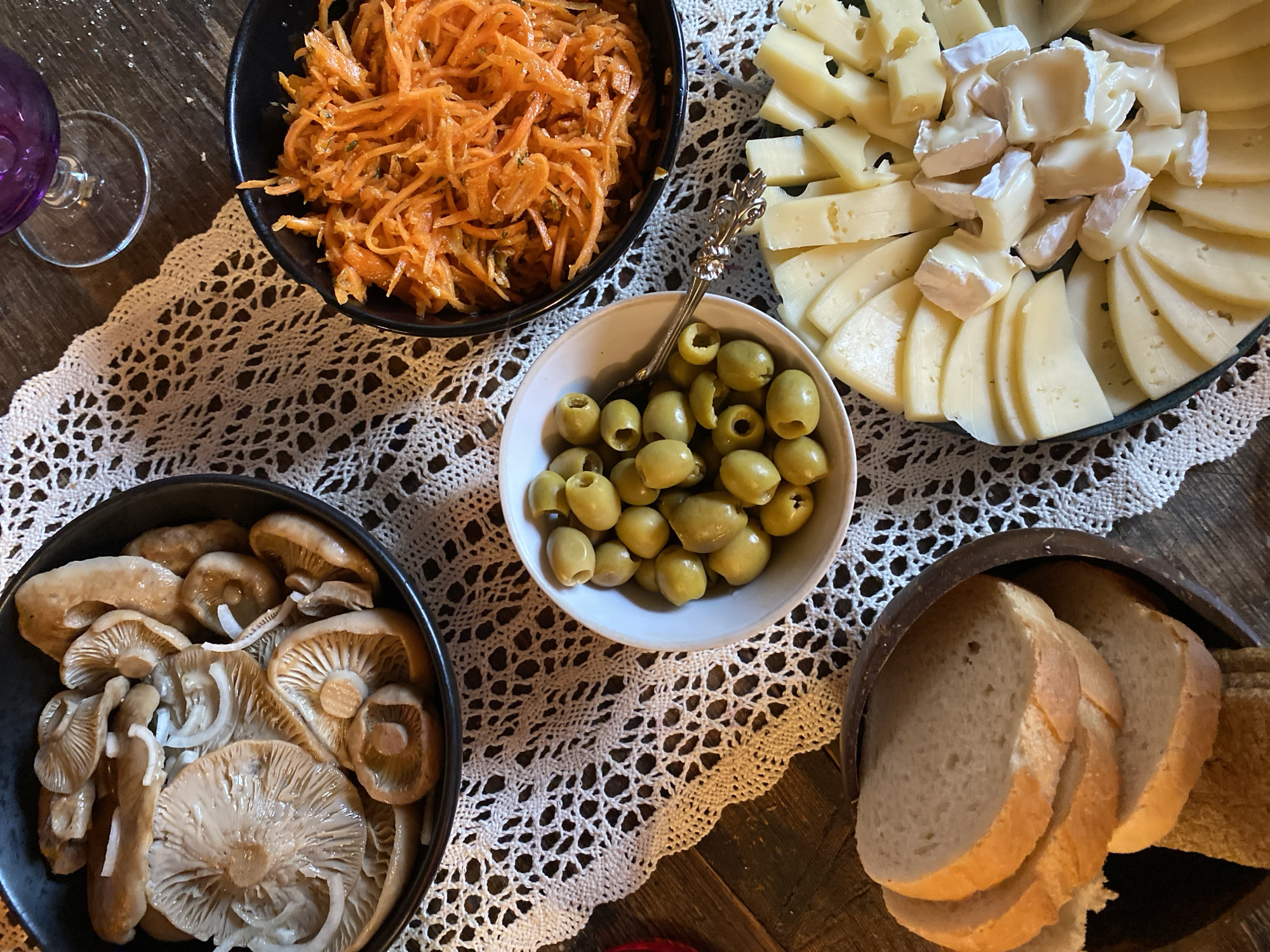 A table is set with an assortment of foods and a drink, including a bowl of pickled mushrooms and onions, a bowl of shredded carrots, a bowl of green olives, sliced bread, and a cheese platter with various types of cheese and a soft white cheese. A glass of deep purple liquid is visible in the upper right corner. The table is covered with a white lace doily. The scene is set outdoors on a wooden surface, likely a table, in Voeikovo, Russia.
