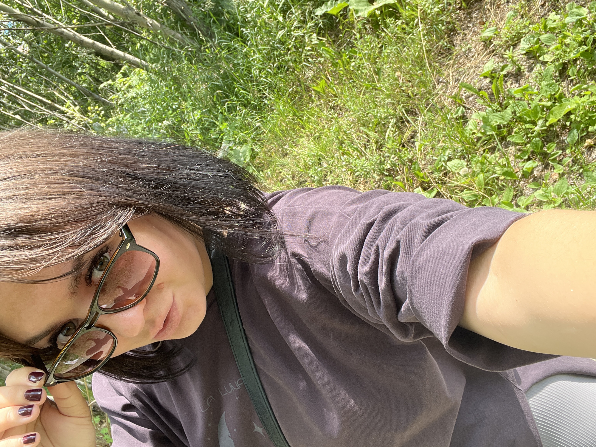A young woman with dark hair and green eyes is taking a selfie outdoors. She is wearing sunglasses and a dark purple t-shirt. Her left hand is adjusting her sunglasses. The background is a blur of green foliage and trees, suggesting she is in a natural setting. The lighting indicates it is daytime.