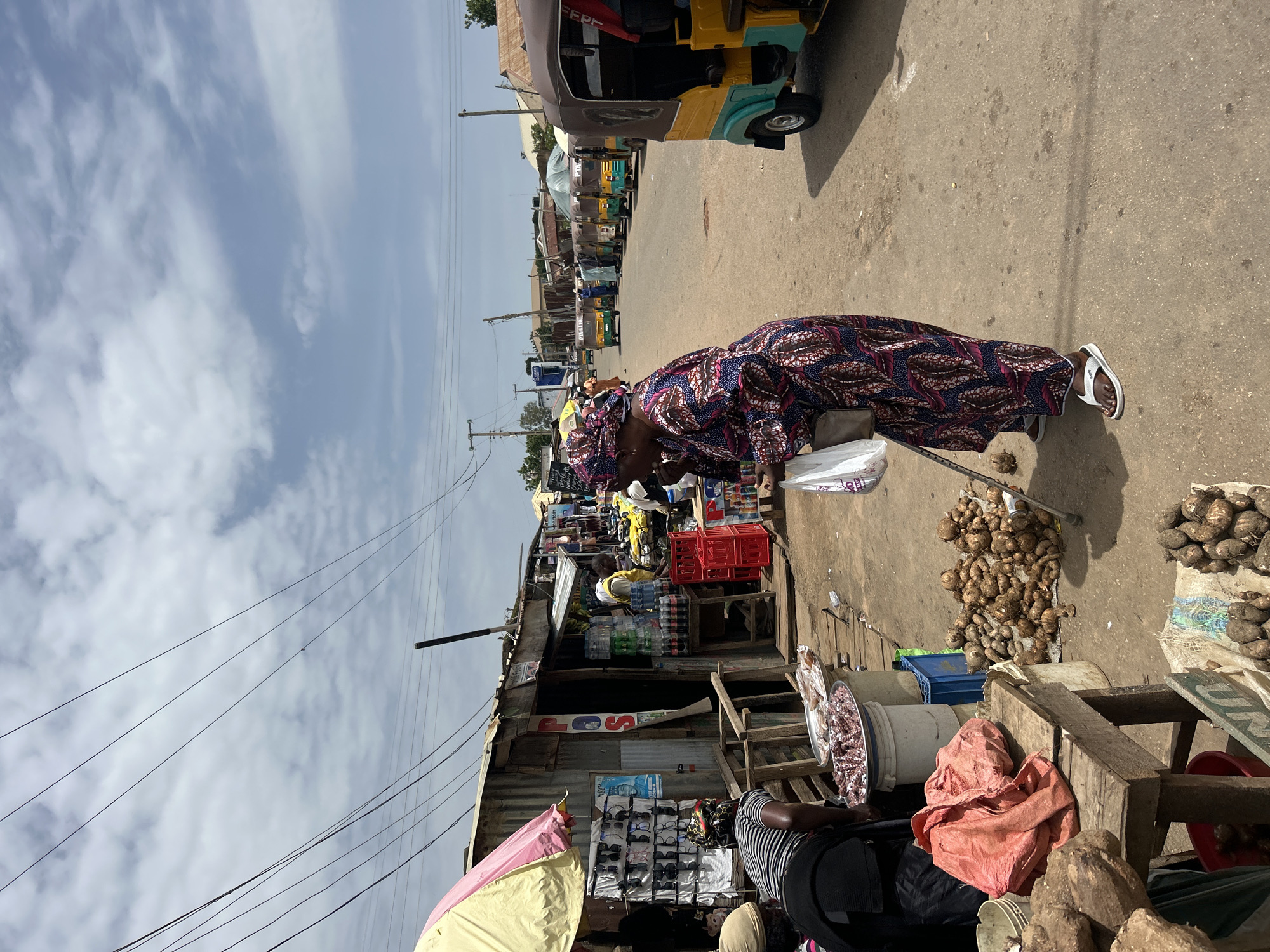 The image captures a bustling market scene in Jos, Nigeria. In the foreground, a woman wearing a vibrantly patterned headwrap and dress is seen leaning on a walking stick. She is dressed in traditional attire, suggesting a cultural context. She appears to be engaged in shopping or attending to a vendor.
The setting is an outdoor marketplace with stalls lining a dirt road. Several "keke napep" (auto-rickshaws) are parked along the road, indicating transportation services are available. The stalls are constructed from corrugated metal and wood, displaying various goods. One stall prominently features sunglasses arranged on a rack, and another has bottles of beverages. Piles of root vegetables, likely yams or taro, are visible on the ground and on display. Bags and fabrics are also present, suggesting a wide array of merchandise.
The weather appears to be partly cloudy with bright sunlight, suggesting it is daytime. The sky has streaks of white clouds against a pale blue backdrop. Overhead, power lines crisscross the frame, a common sight in urban and semi-urban areas.
The overall atmosphere is that of an active commercial hub, with people engaged in their daily activities. The image conveys a sense of everyday life in a Nigerian market. Some text is visible on signage and product packaging, but it is not clearly legible to extract specific information.