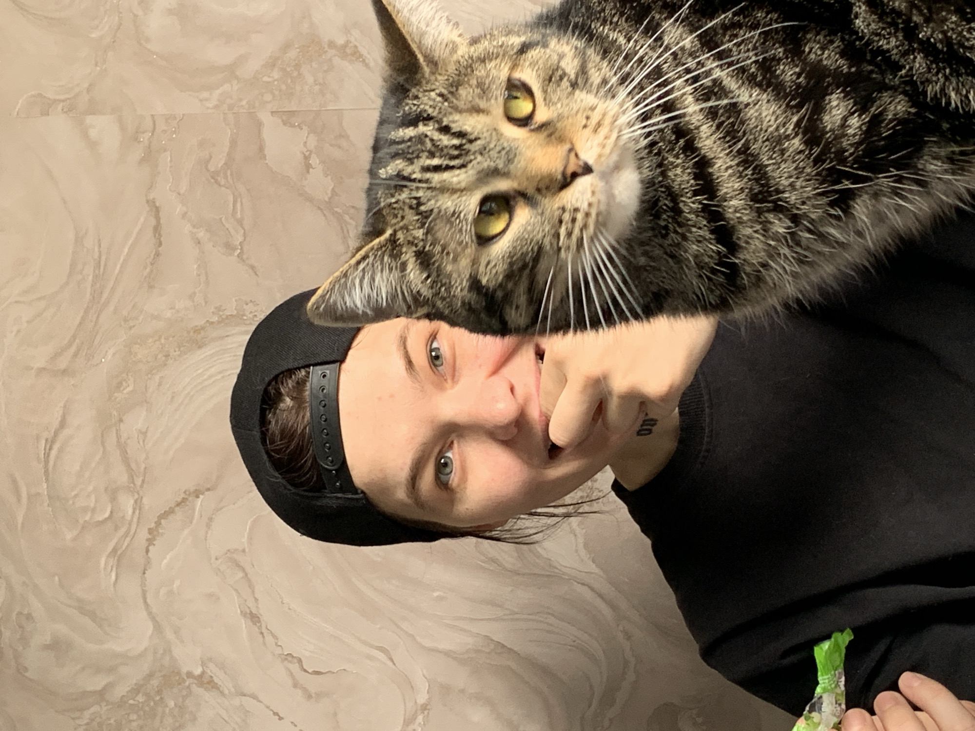 In a close-up, waist-up shot, a person wearing a black baseball cap and a black sweatshirt is partially obscured by a tabby cat. The person's face is turned to the left, with their right hand near their mouth, and they are smiling. Their left hand is visible in the lower left corner holding a green candy wrapper. The cat, positioned to the right of the person, fills a significant portion of the frame, with its head and chest in focus. The background is a textured, light-colored wall with a marble-like pattern.