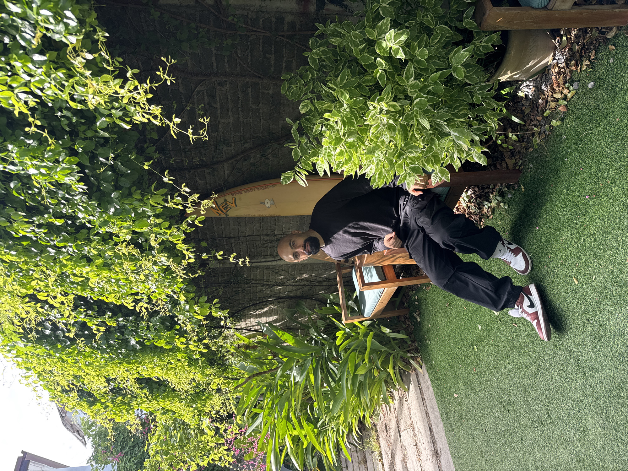 In a courtyard in Ko Samui District, Thailand, a bald man with a beard sits on a wooden chair with a teal cushion. He wears a black long-sleeved shirt and black pants. On his feet are maroon and white sneakers. A tan surfboard with orange trim stands upright against a brick wall behind him. Lush green plants, some with variegated leaves, surround the man and the chair. The ground is covered in artificial grass, with a stone pathway visible to the left. Sunlight filters through the dense green foliage overhead, creating dappled shadows.
