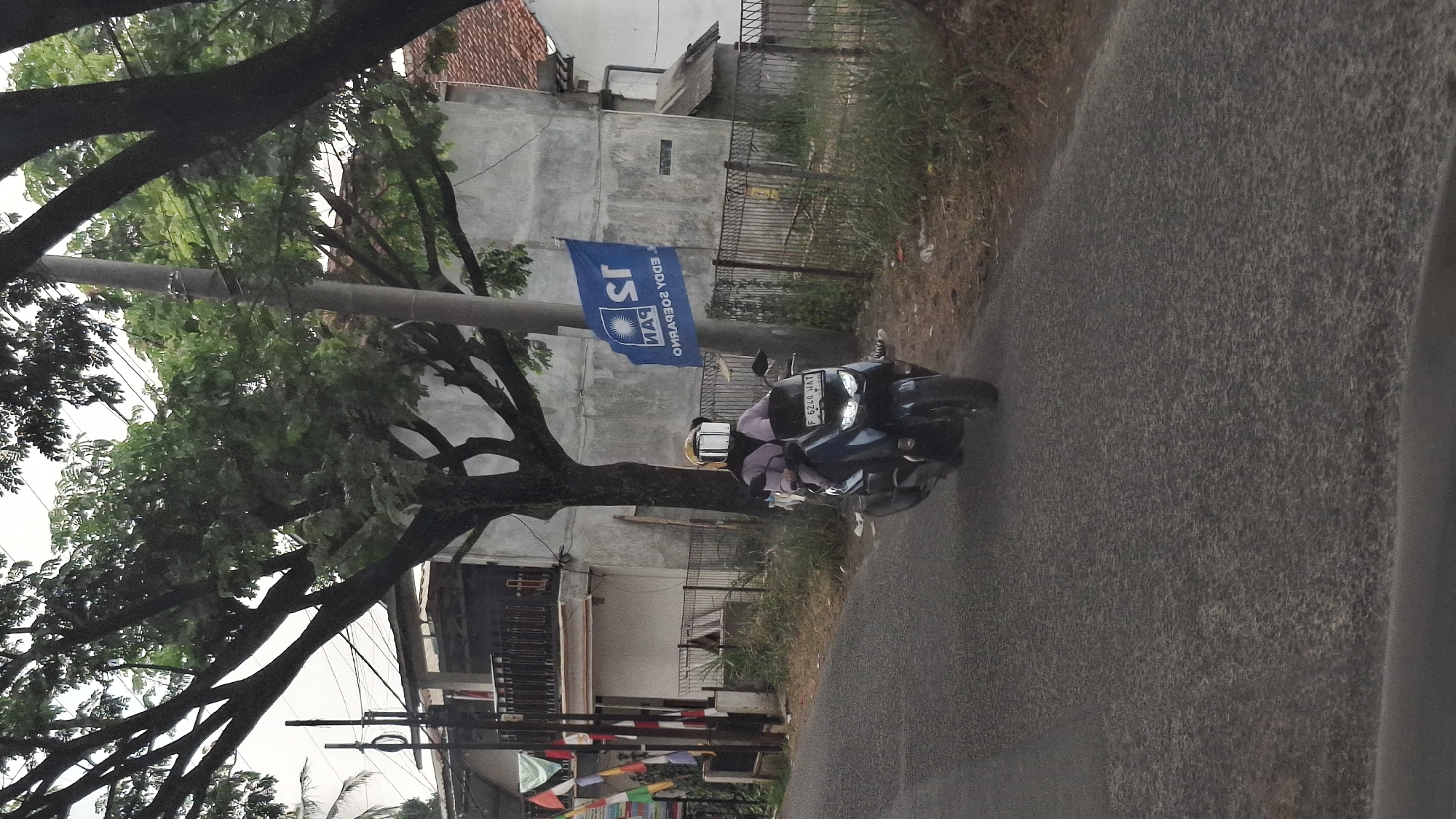A person is riding a dark-colored motorcycle down a paved road. The rider is wearing a purple shirt, a dark headscarf, and a yellow helmet with a white, rectangular visor. The motorcycle has a license plate that reads "F 6240 HAT". A blue flag with white lettering and a sun-like logo is attached to a pole on the right side of the road, behind the motorcycle. The flag has text that appears to be "PAN" and "EDDY SOEPARNO". The environment is leafy with trees on the left and right sides of the road. In the background, there is a light-colored building with a red-tiled roof and a fence. The image appears to be taken in Cibinong Hilir, Indonesia, based on the information provided.