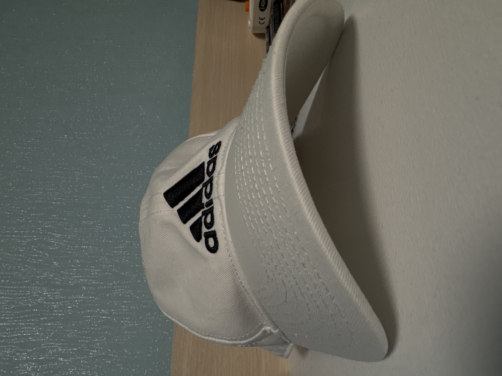 A white baseball cap with the Adidas logo is resting on a light brown or beige surface next to a light blue wall and a white wall. The cap is oriented sideways, with the bill facing right. The logo, consisting of three black stripes and the Adidas wordmark, is visible on the side of the cap facing the viewer. A small object with text is visible behind the top of the cap. The lighting suggests an indoor environment. The city and specific location are unknown.