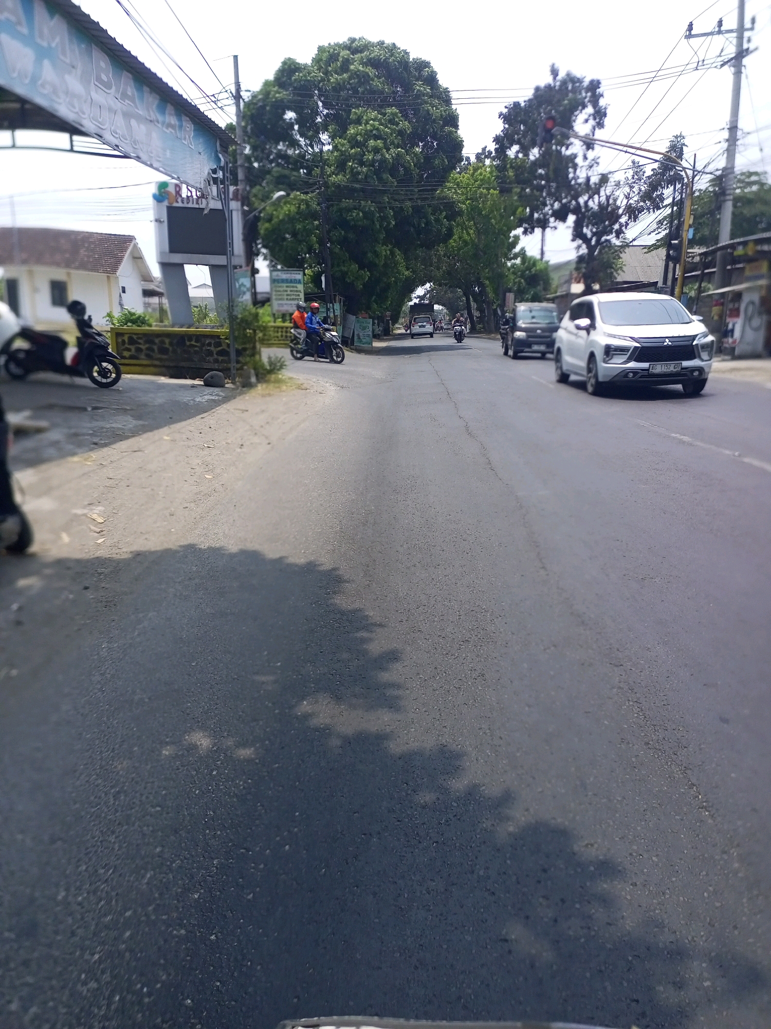 In Ngasem, Indonesia, a street scene unfolds with a white SUV in the foreground. The SUV, license plate AG 1152 GII, is traveling on an asphalt road. In the background, a motorcycle with two individuals wearing orange and blue helmets is visible. Further down the road, another motorcycle is parked to the left, and a white house with a brown roof is situated behind a low stone wall. Overhead, power lines crisscross the sky, and lush green trees arch over the road, creating a natural canopy. A banner is displayed on the left side of the street, partially obscured by power lines. The scene is captured during daylight hours, with shadows cast on the road by trees and possibly other objects.