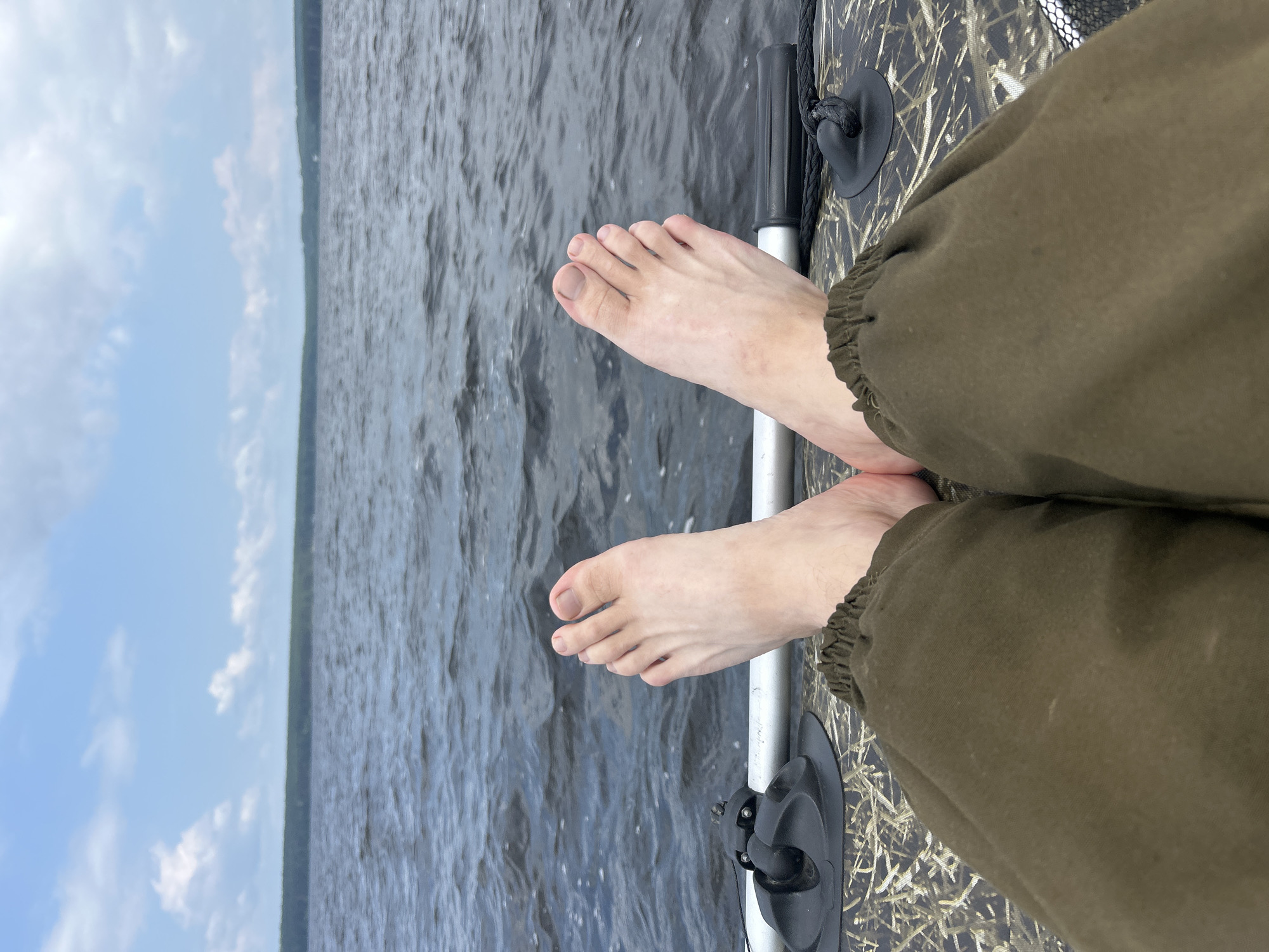 The image depicts a first-person perspective of someone sitting on the edge of what appears to be a boat or raft on a body of water. The person's bare feet are visible, resting on the textured surface of the vessel. Their legs are covered by dark green pants. In the background, a calm, dark blue body of water stretches towards a distant treeline under a sky with scattered white clouds. The scene is likely set on a lake or a wide river. The image is taken in Zabegaevo, Russia.