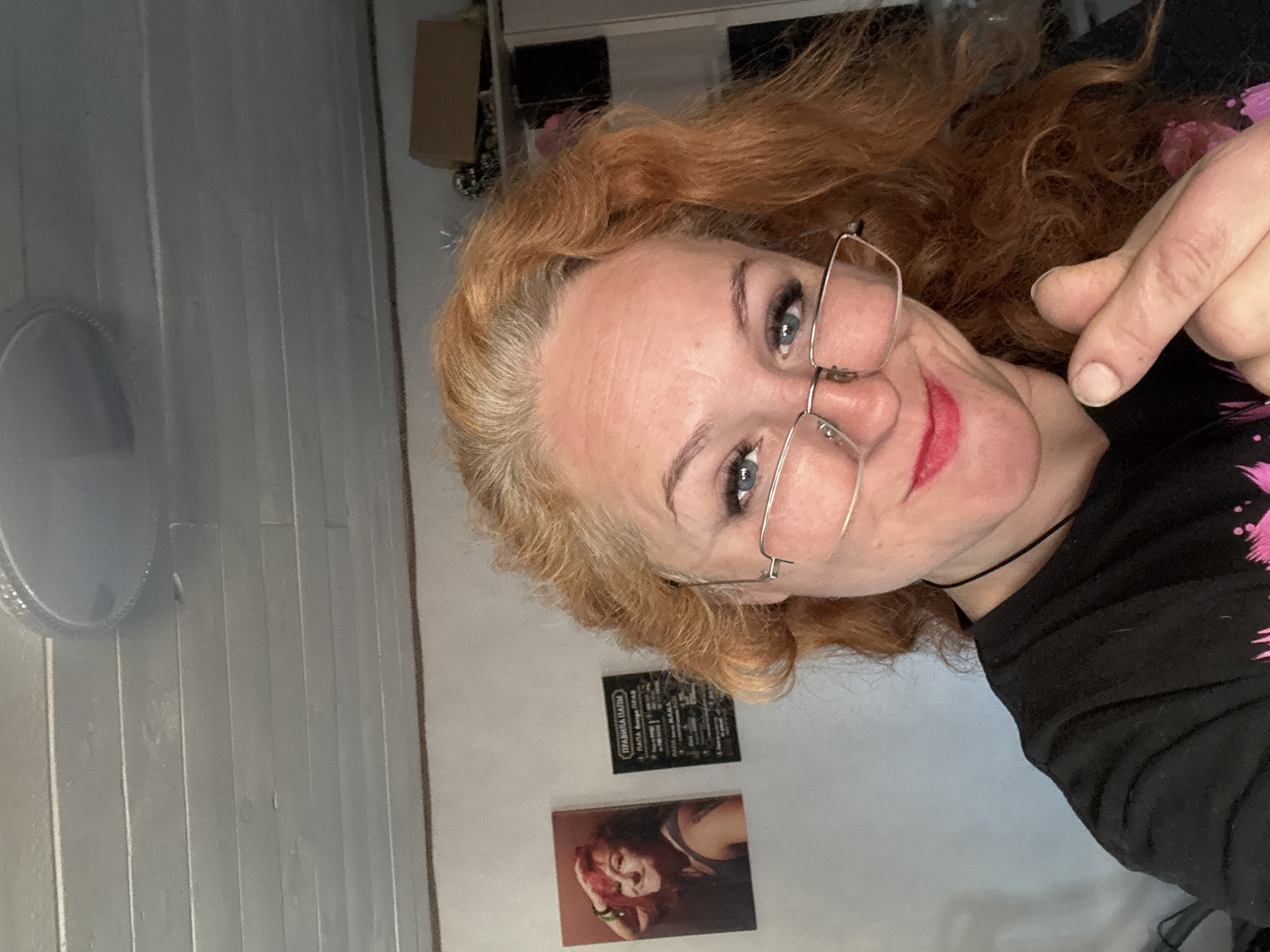 A selfie depicts a woman with fair skin, blue eyes, and red, curly hair, wearing glasses and dark clothing. She is pointing with her right hand towards the viewer. The background includes a white paneled wall with a round, white object (possibly a fan) on the left. Two small, framed artworks are visible on the wall behind her, one appearing to be a print of another woman and the other a framed graphic. The location is suggested to be in the city of Chaikovsky, Russia.